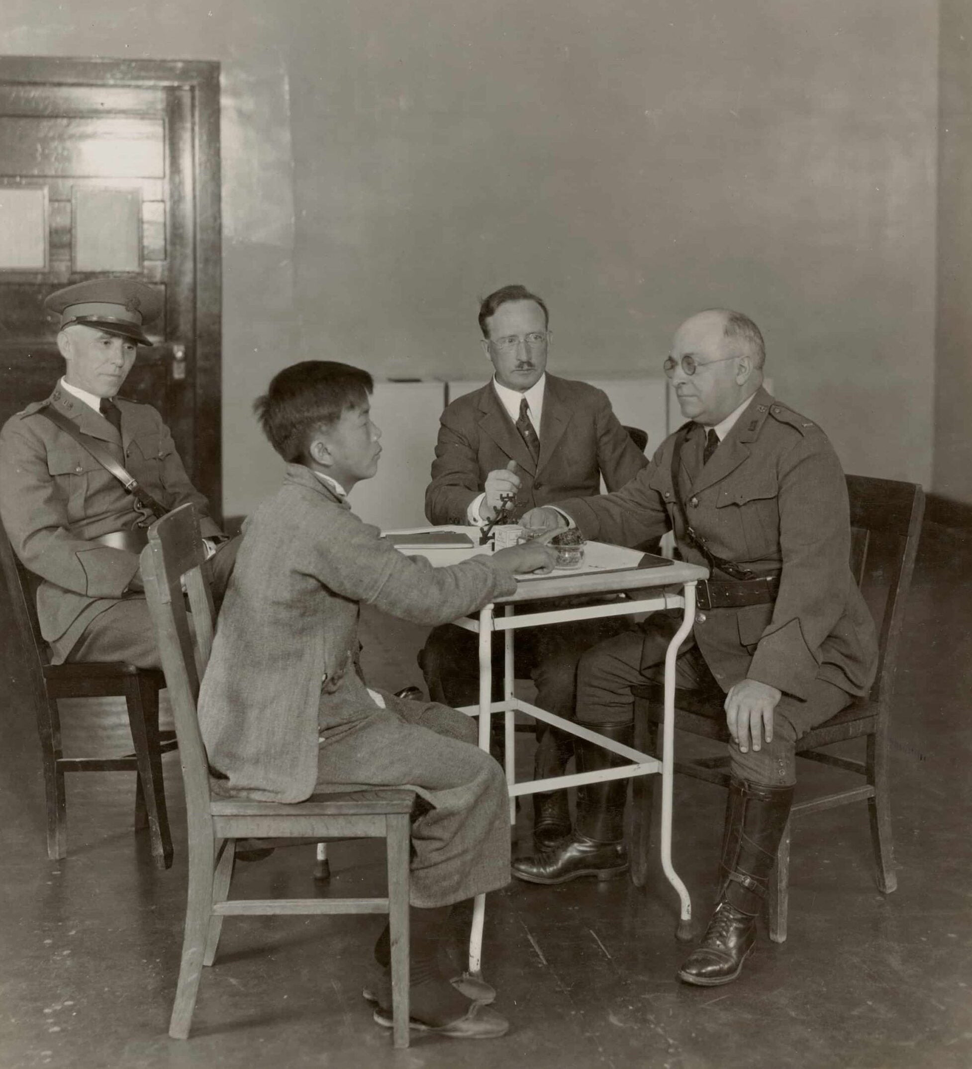Two white men in prison guard outfits and a white man in a suit sits with a Chinese boy in a suit and interrogates him.
