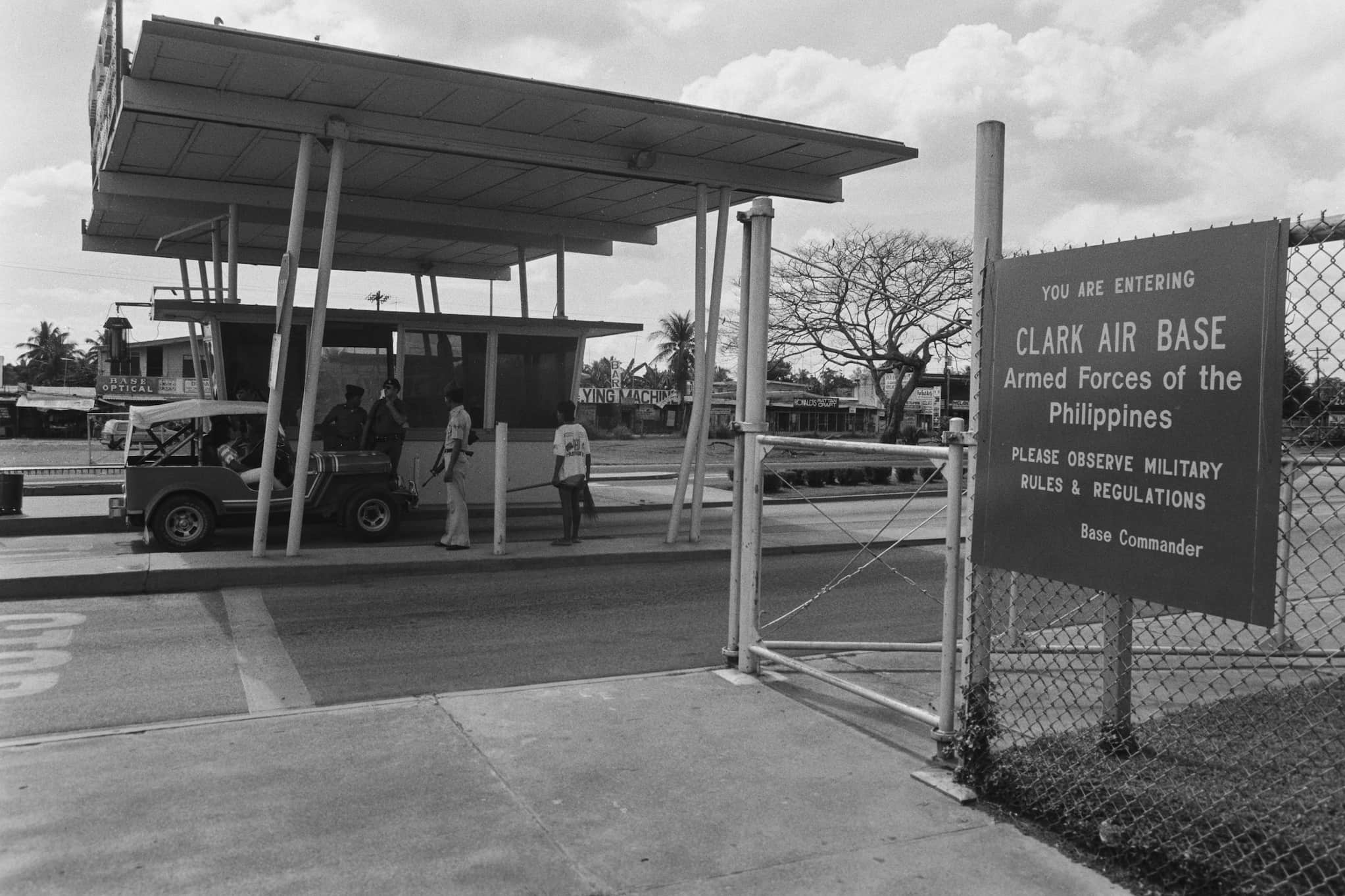 Black and white photo of the Clark Air Base entrance gate. A jeep is stopped at the security booth with three men in uniform.