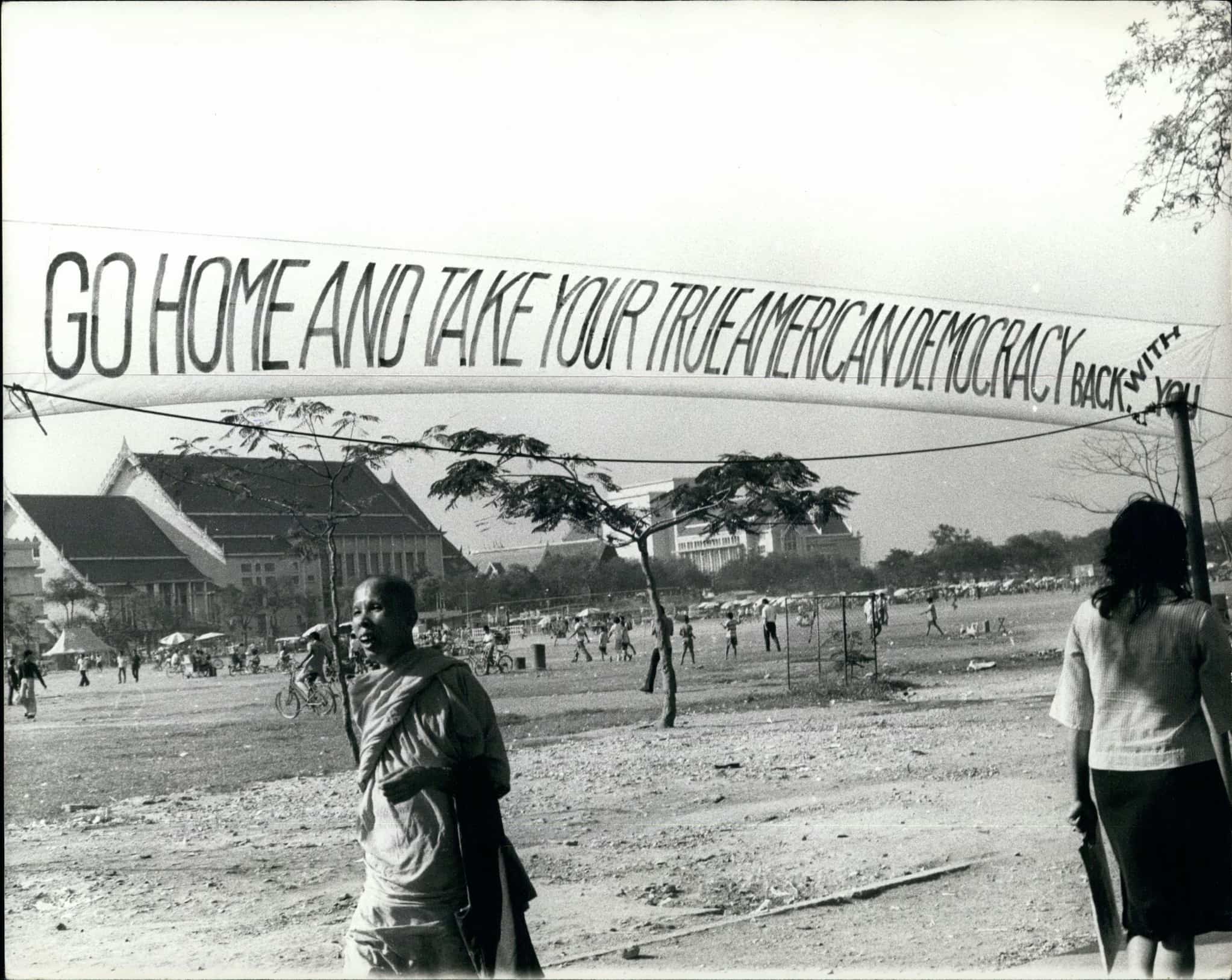 Large banner with text "Go Home and Take Your True American Democracy Back With You" hangs in front of large field crowded with people.