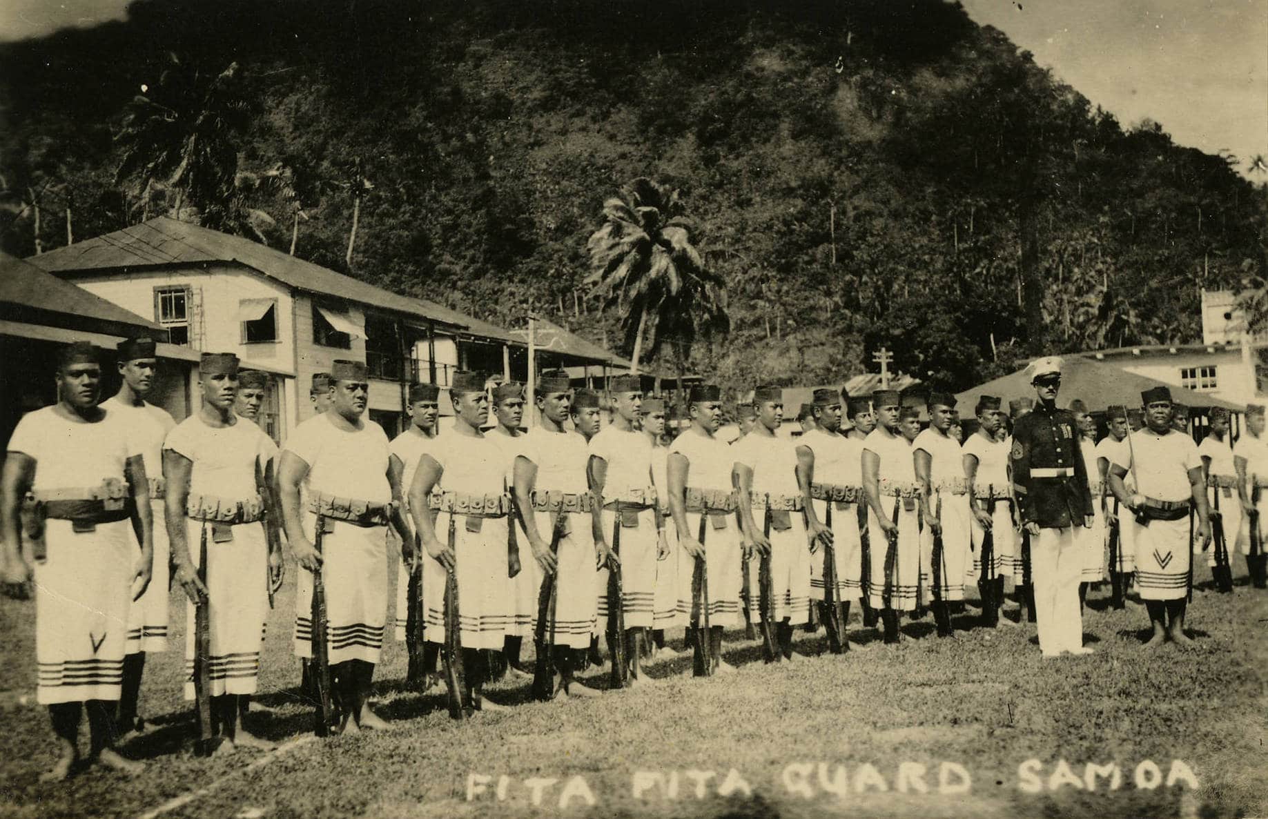 Fita Fita Guards with rifles stand in organized line behind one US soldier. Buildings and a steep, vegetation-covered mountain stand behind them.