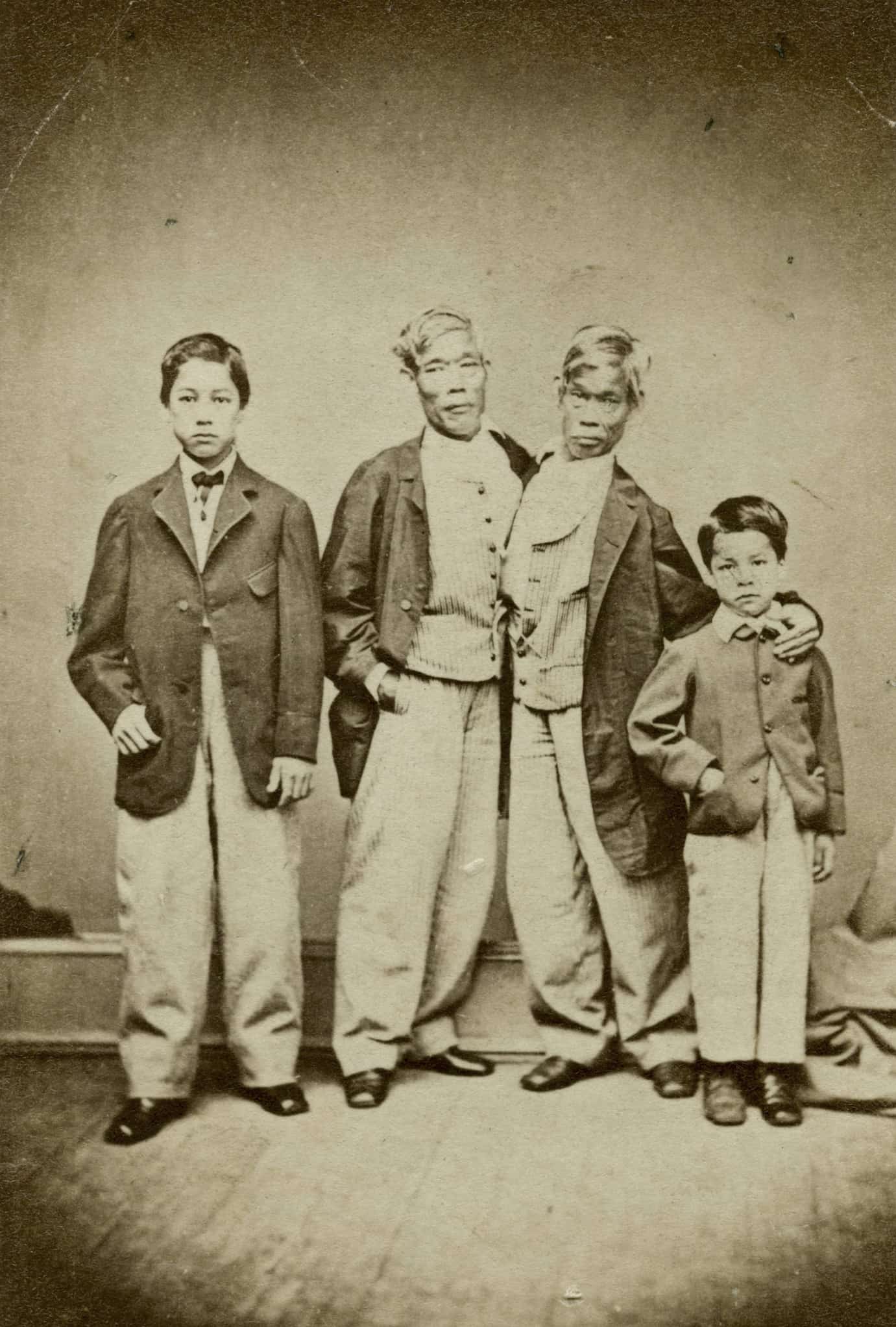 Eng and Chang Bunker, conjoined twins from Thailand, dressed in trousers, vests, and jackets stand with their two sons in a family portrait.