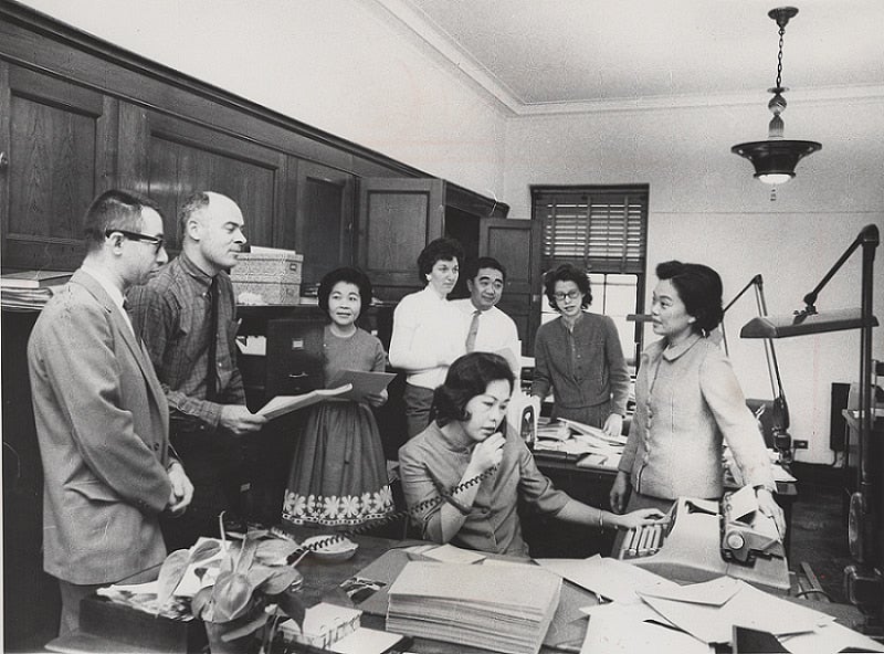 Patsy Mink stands near typewriter as she addresses her office staff. Her staff, men and women seated and standing, gather in half circle around her.
