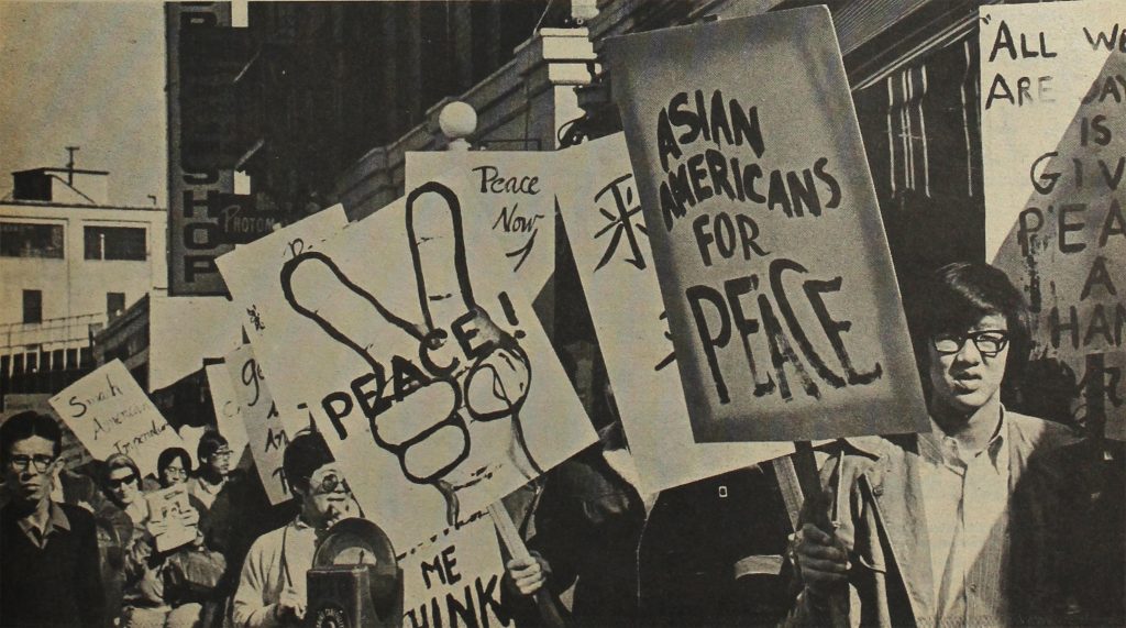 Crowd of protestors with large signs march down the sidewalk of an urban street. Man in glasses holds large sign reading "Asian Americans for Peace."