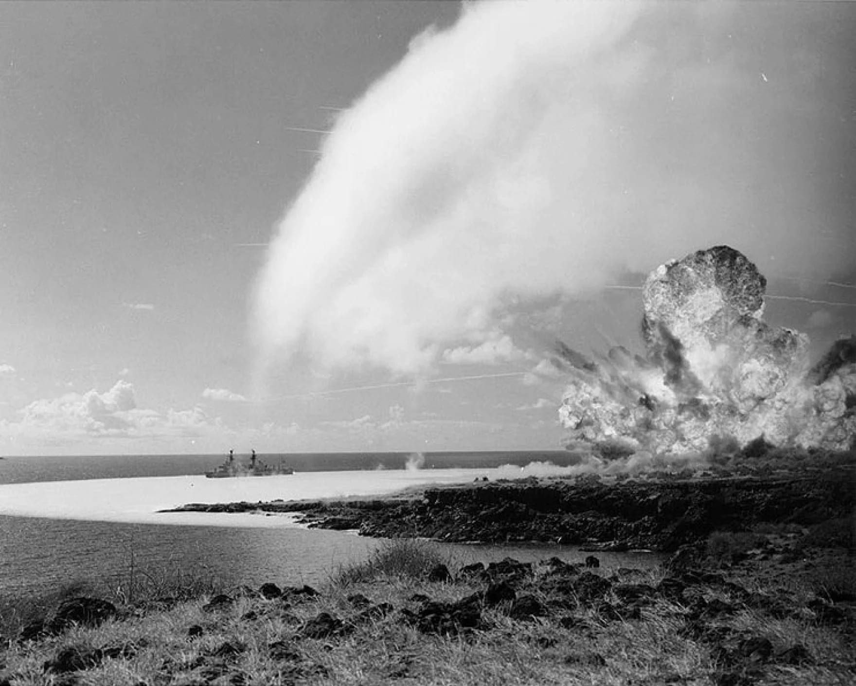 Clouds from explosives test on the rocky shore of Kaho'olawe Island billow towards sky. A large naval ship is in the water left of explosion.