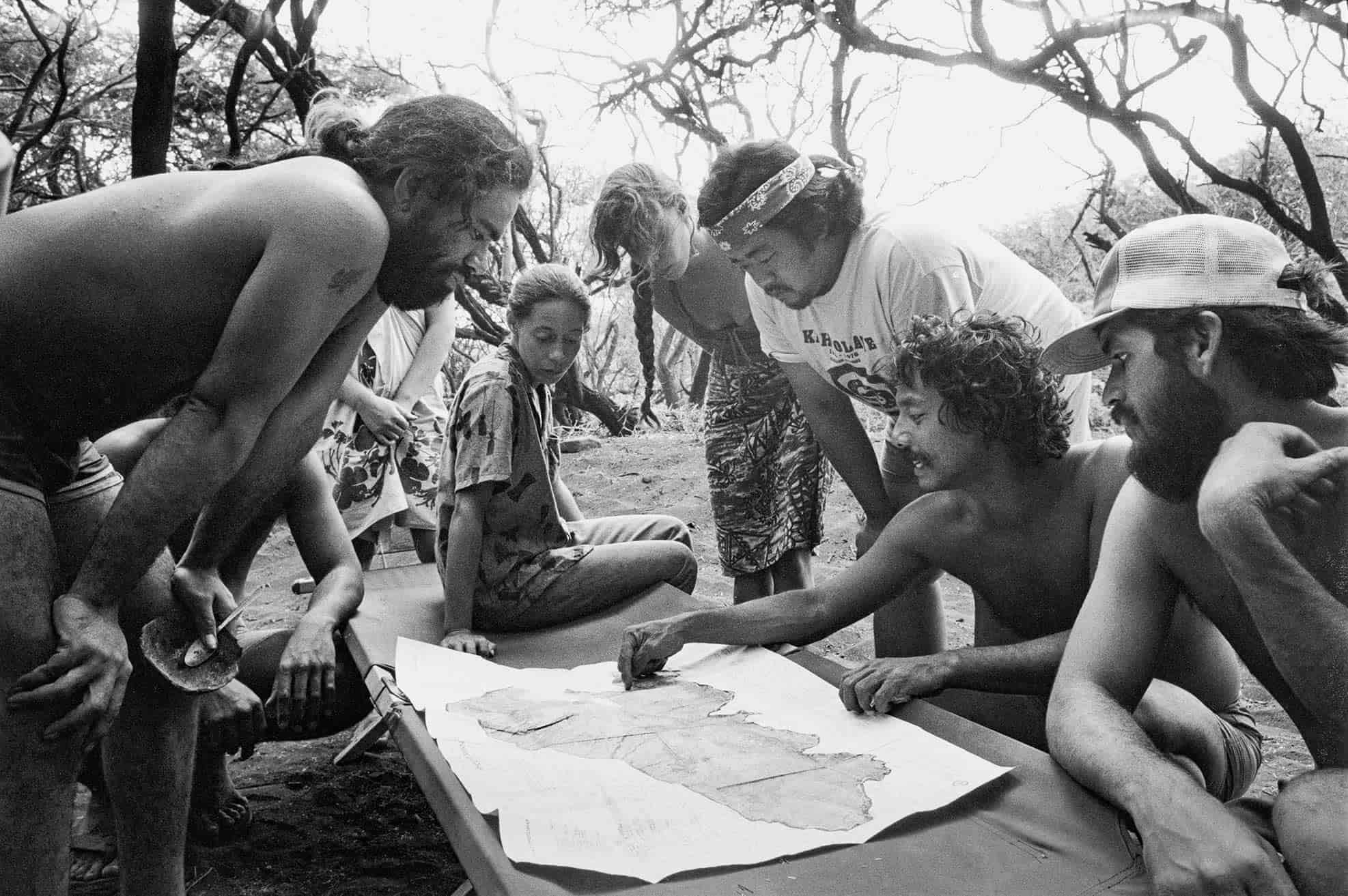 Group of young people, dressed in warm weather clothing, surround outdoor cot with map of Kahoʻolawe between them. Denuded trees in the background.