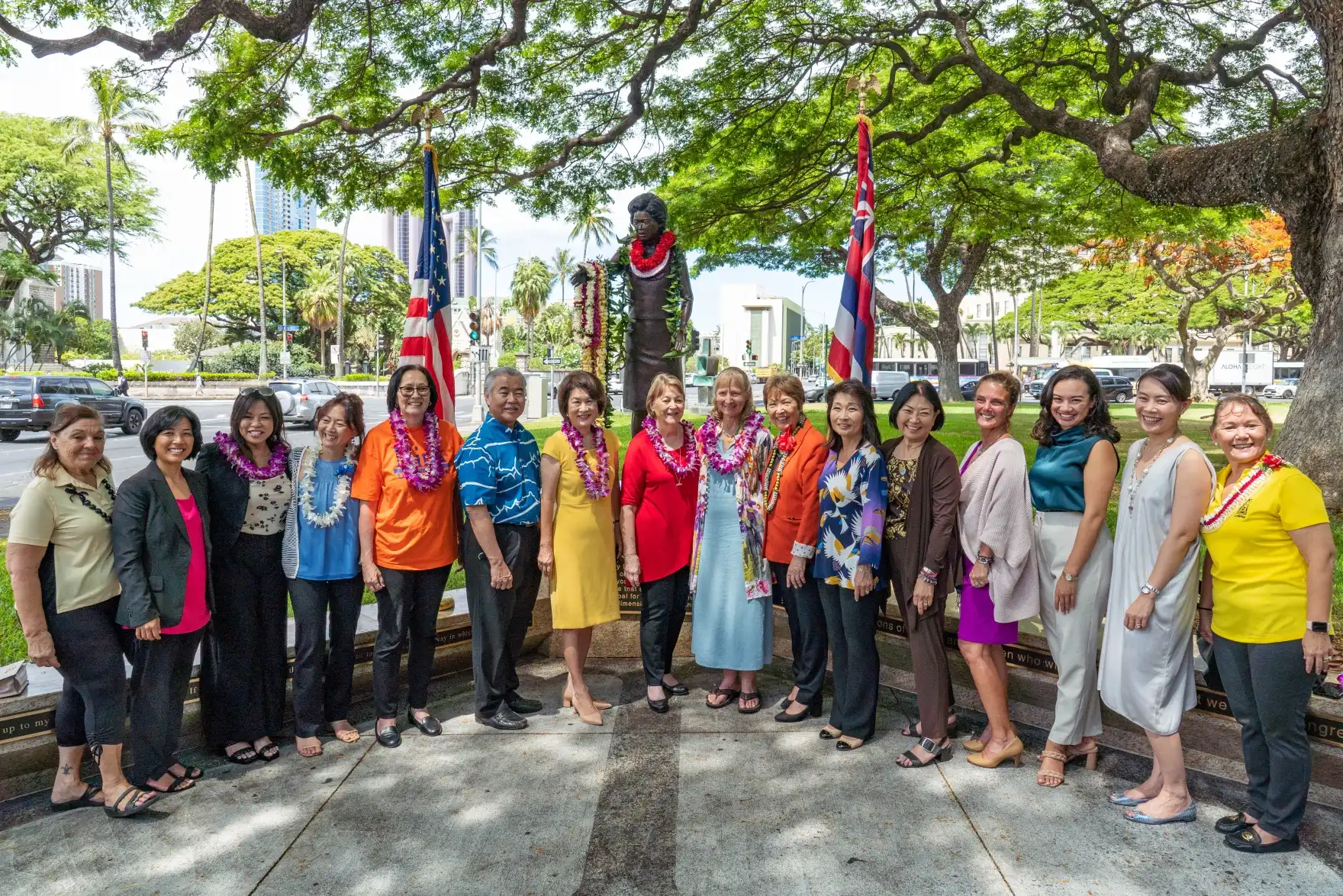 Group in professional attire and lei stand before outdoor statue of Patsy Mink. The flags of the U.S. and Hawaiʻi are raised on either side of statue.