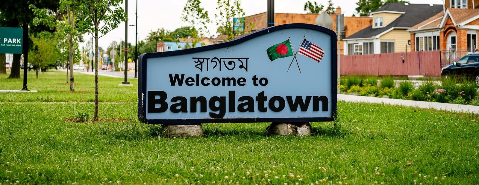 A sign with concrete footings placed on a lawn that reads "Welcome to Banglatown". Images of a Bengali flag and an American flag are next to the text.