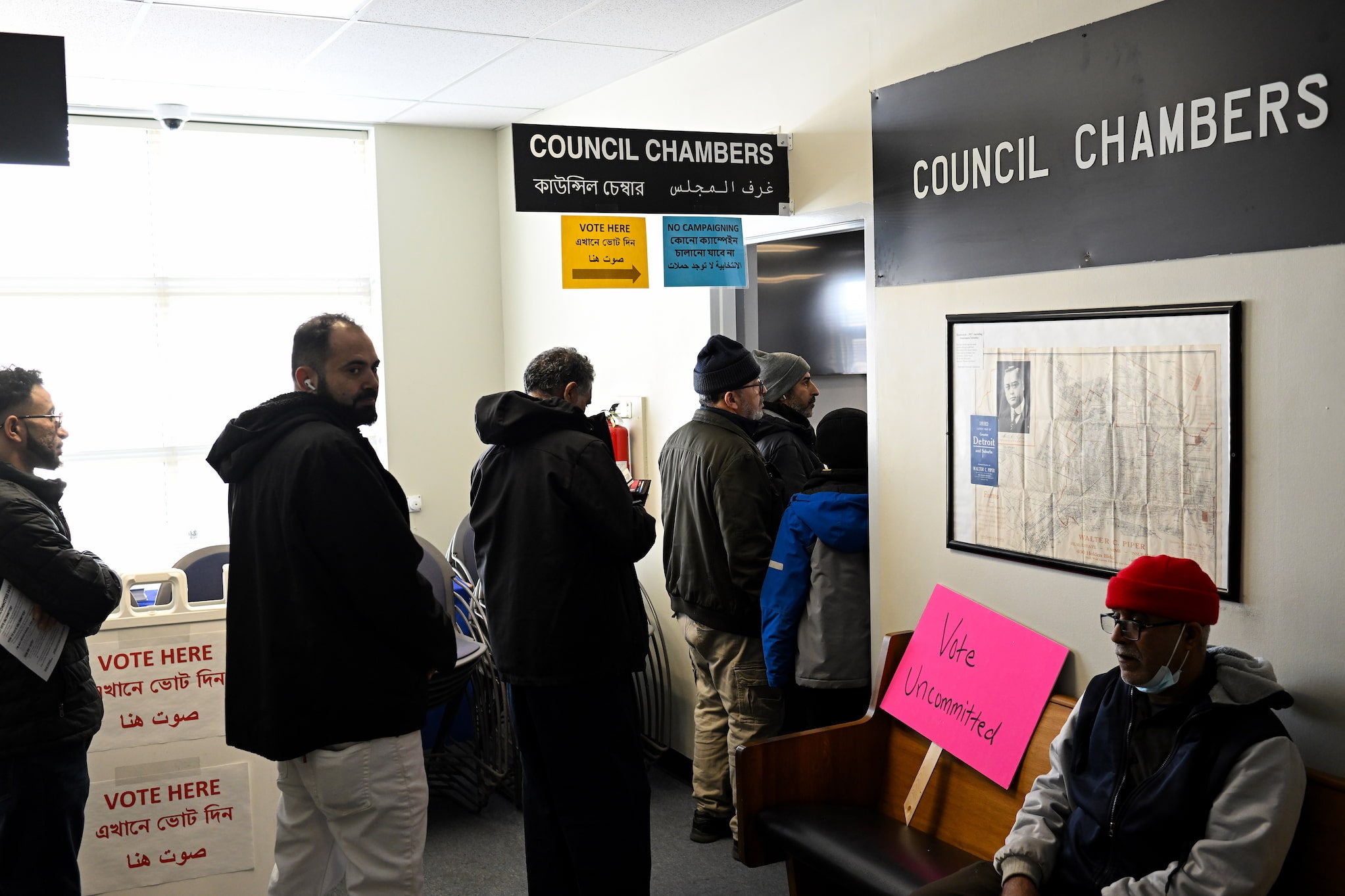 Arab and Muslim Americans wait in line at a polling station in the council chambers. A pink sign sits on a bench that reads "Vote uncommitted".