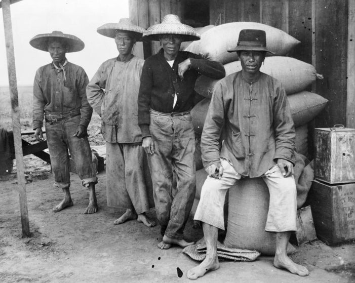 Four barefoot Chinese field hands wearing a mix of shanku and western clothing, with straw hats loiter in front of stacked sacks of harvested grains.