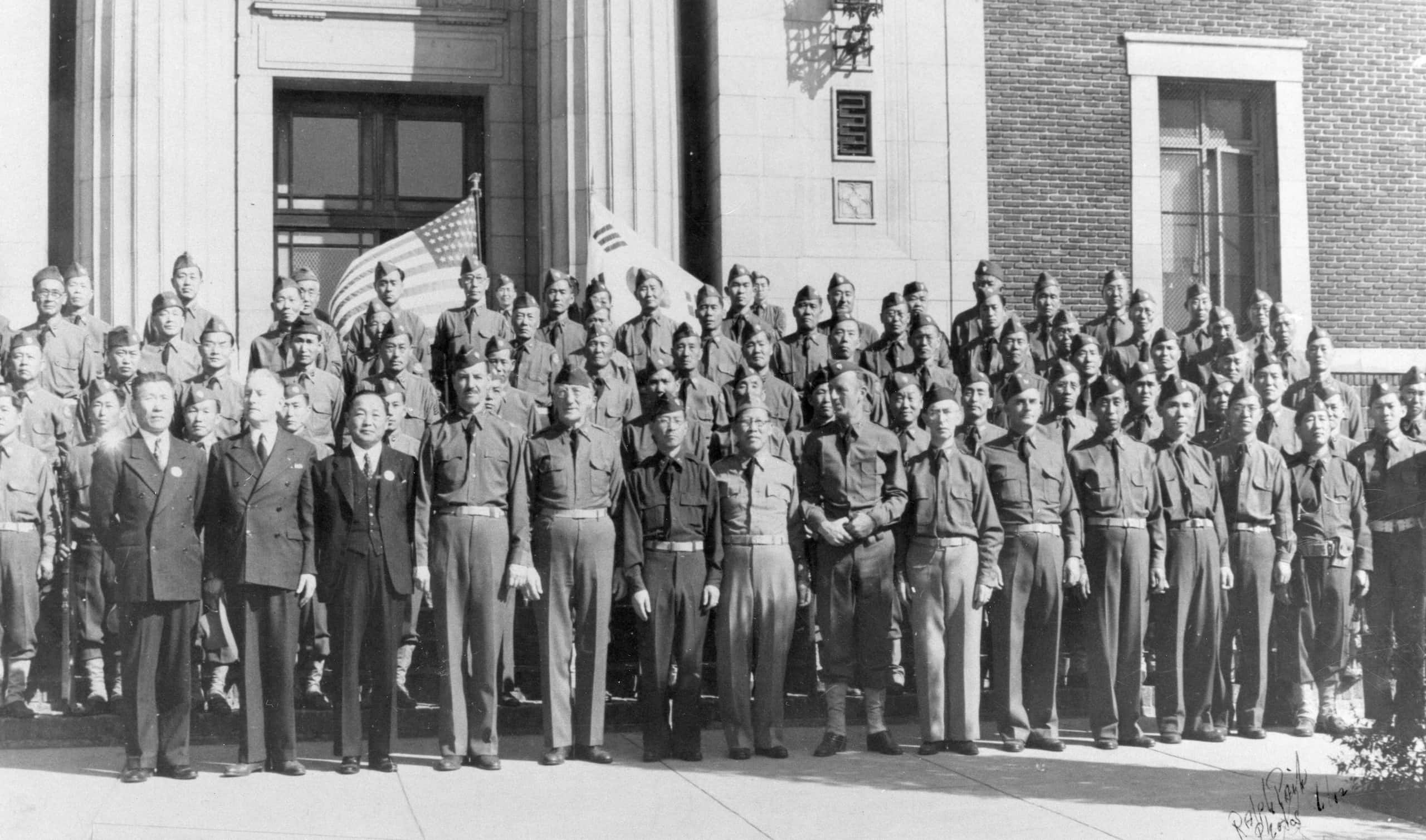 Uniformed Korean American men stand in rows outside of building. Flags of the U.S. and South Korea are seen behind last row of men.