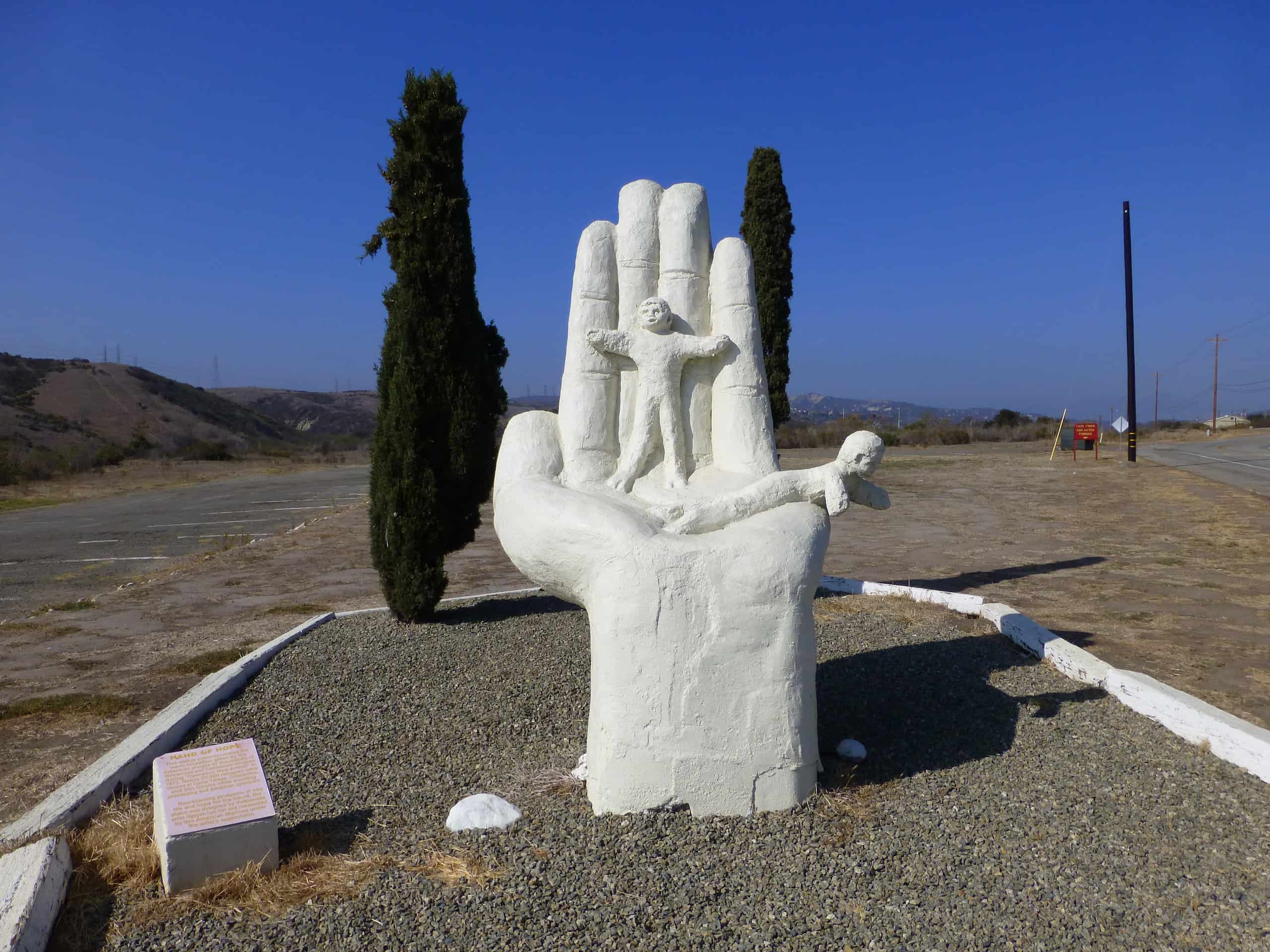 Large white cement statue of an open palm with a little girl standing along the fingers while a little boy lays down on the palm.