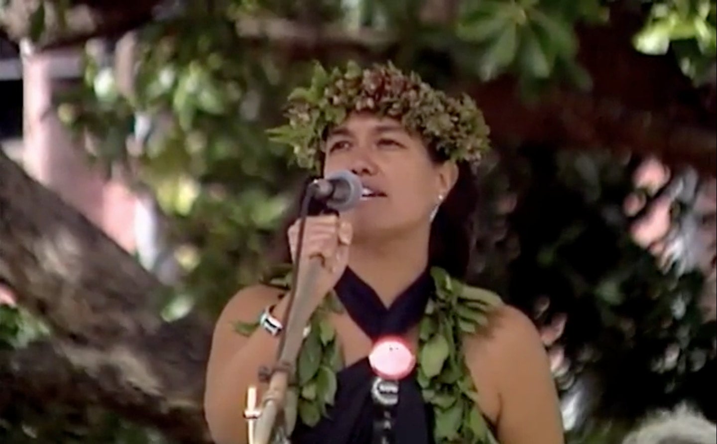 Haunani-Kay Trask, a woman with long, dark hair wearing lei poʻo, speaks passionately into microphone at outdoor event.