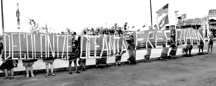 Twelve people, standing in a row, carry four, large banners bearing the words "Decolonize Oceania, Free Guam" at outdoor festival in Guam.