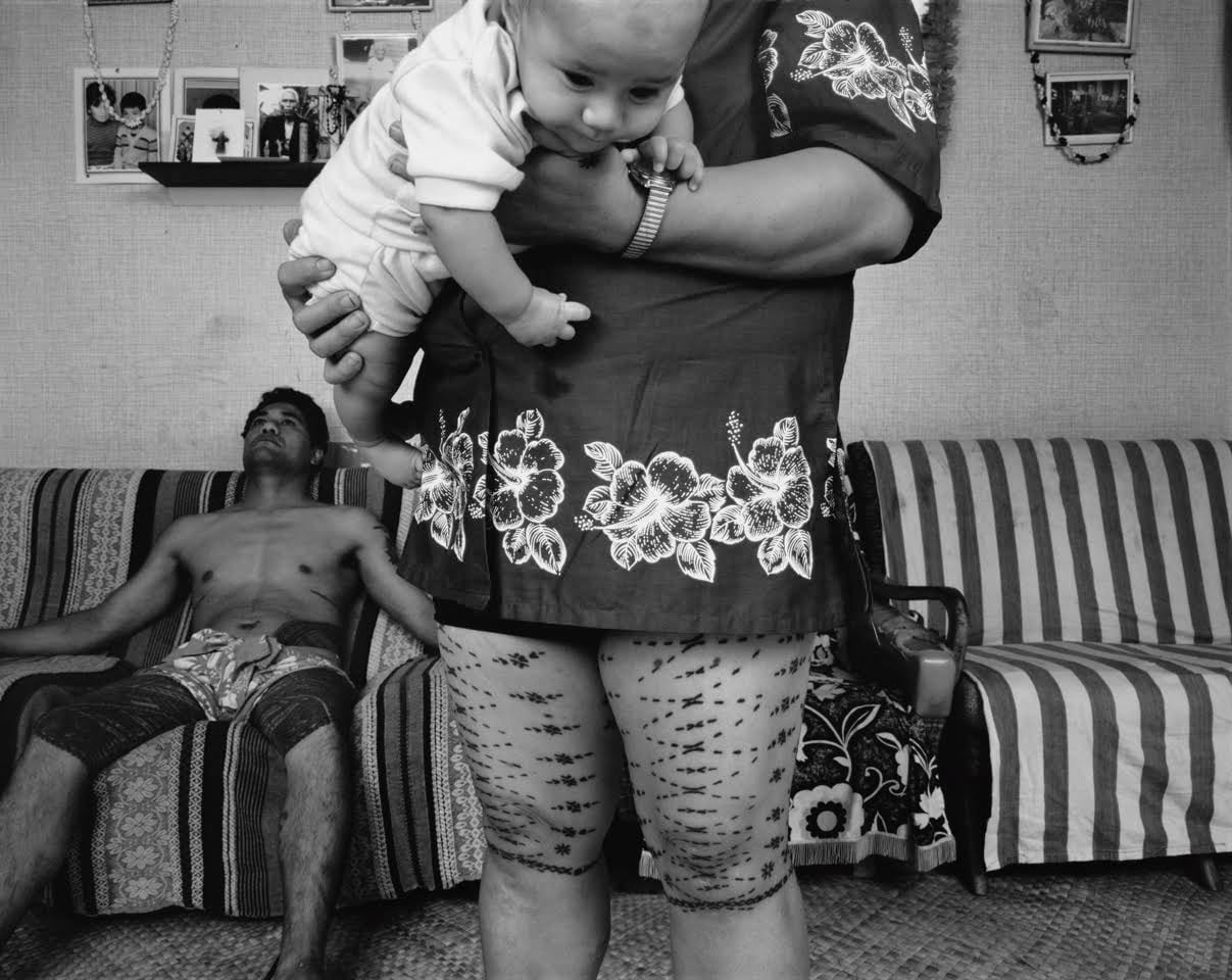A Samoan woman, carrying a baby, stands in front of a couch with a shirtless man sitting on the left. The malu on her legs are featured.