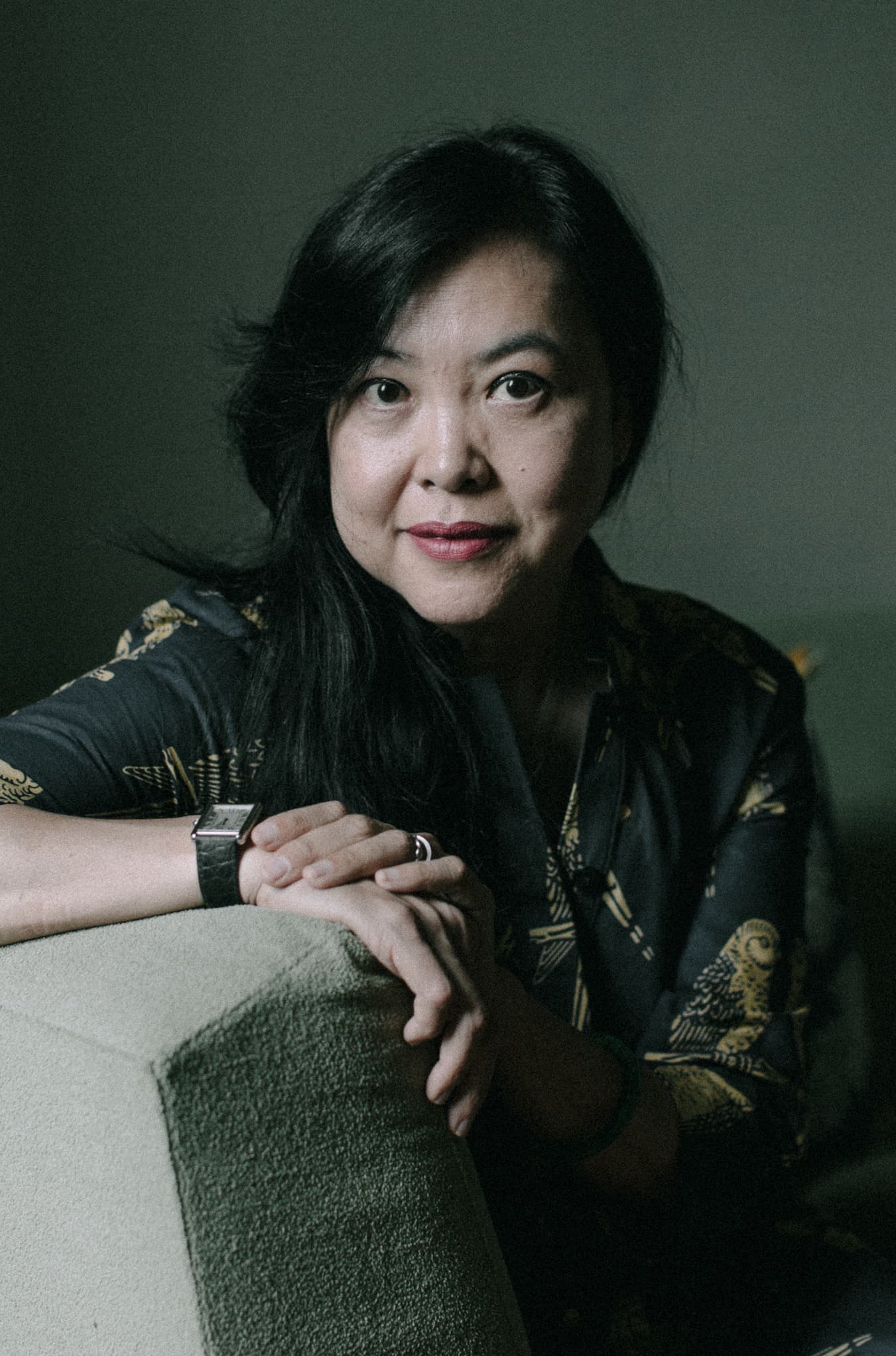 Headshot of Vietnamese American Monique Truong who is leaning on a padded chair and wearing a dark, patterned blouse.