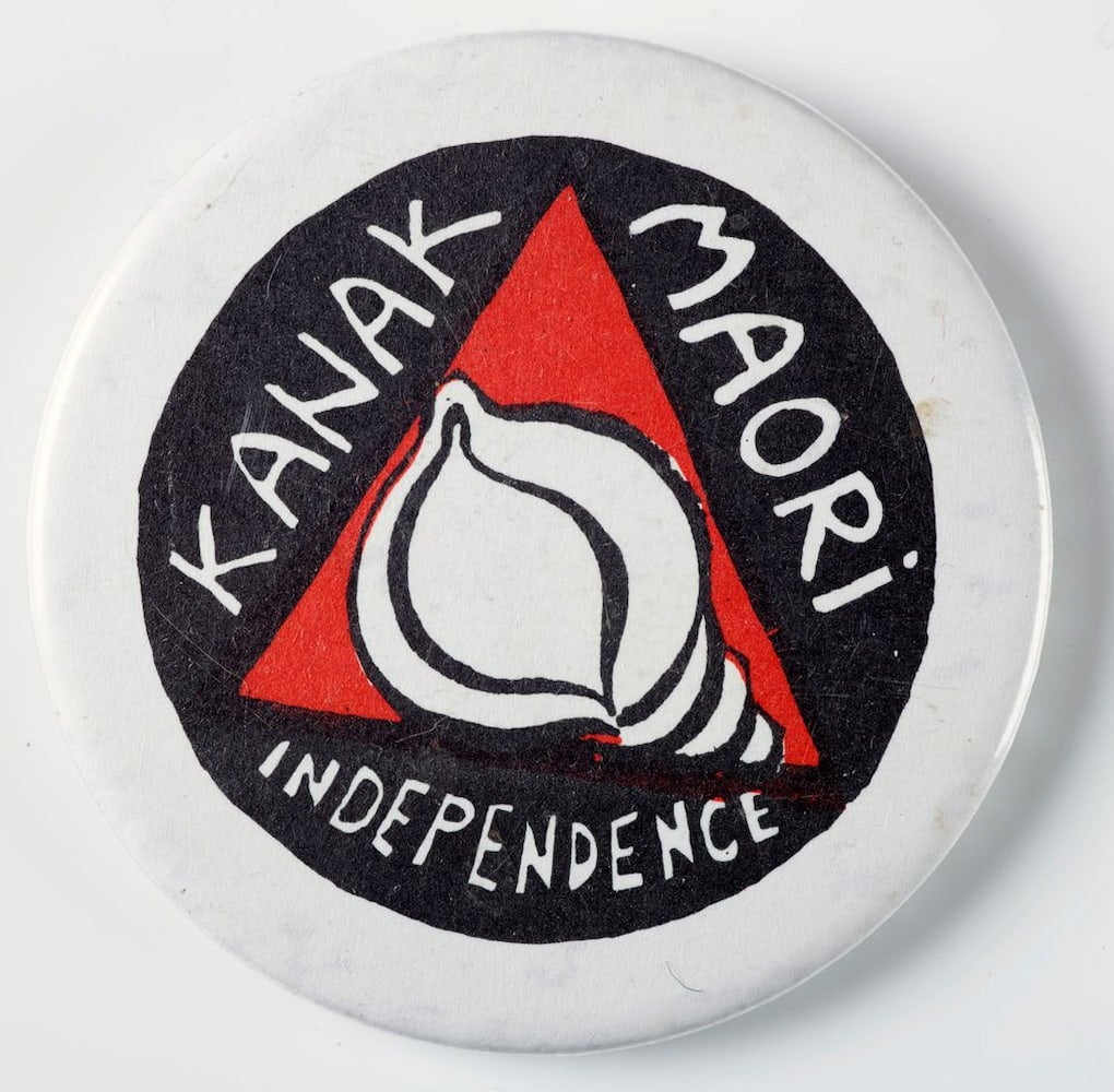 A white, circular badge features a red triangle with a conch shell in the center. Surrounding it is text that reads, "Kanak Maori Independence."