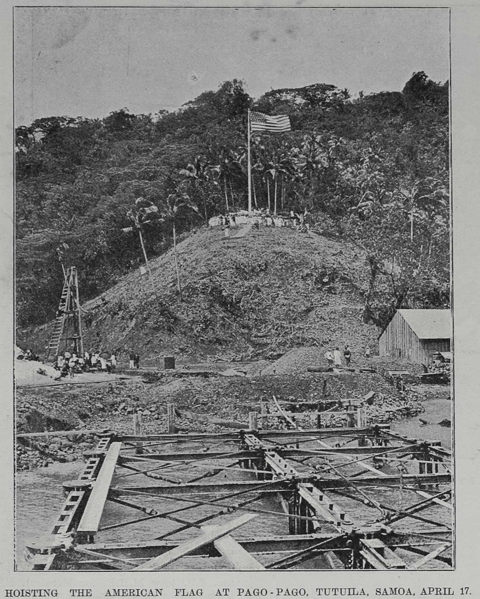 On top of a hill surrounded by palm trees and a forest, an American flag flies on a flagpole. At the base of the hill are people and construction.