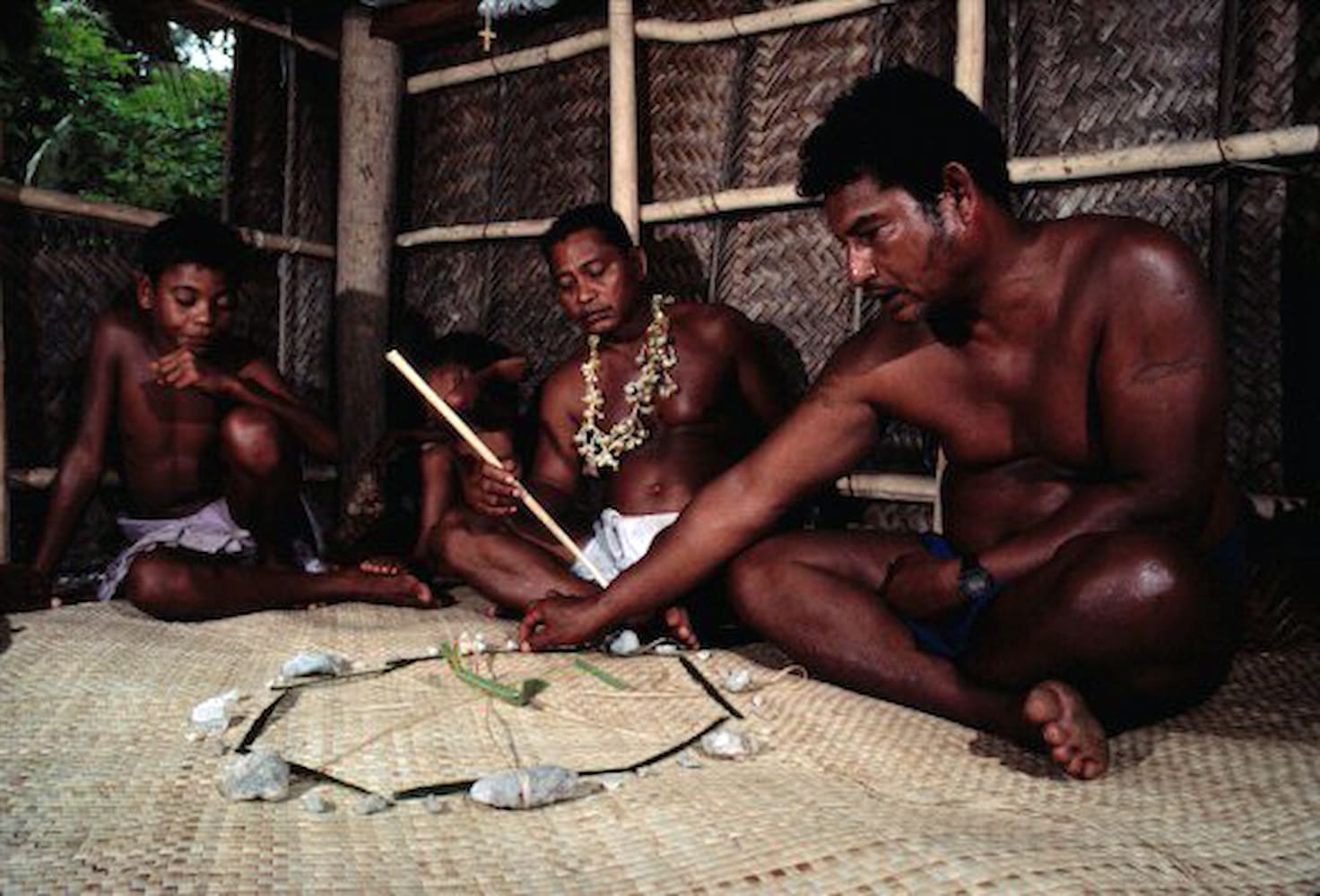 Mau Piailug sits cross-legged and points to a diagram on woven mat. He teaches navigation to two kids and a man seated next to him.