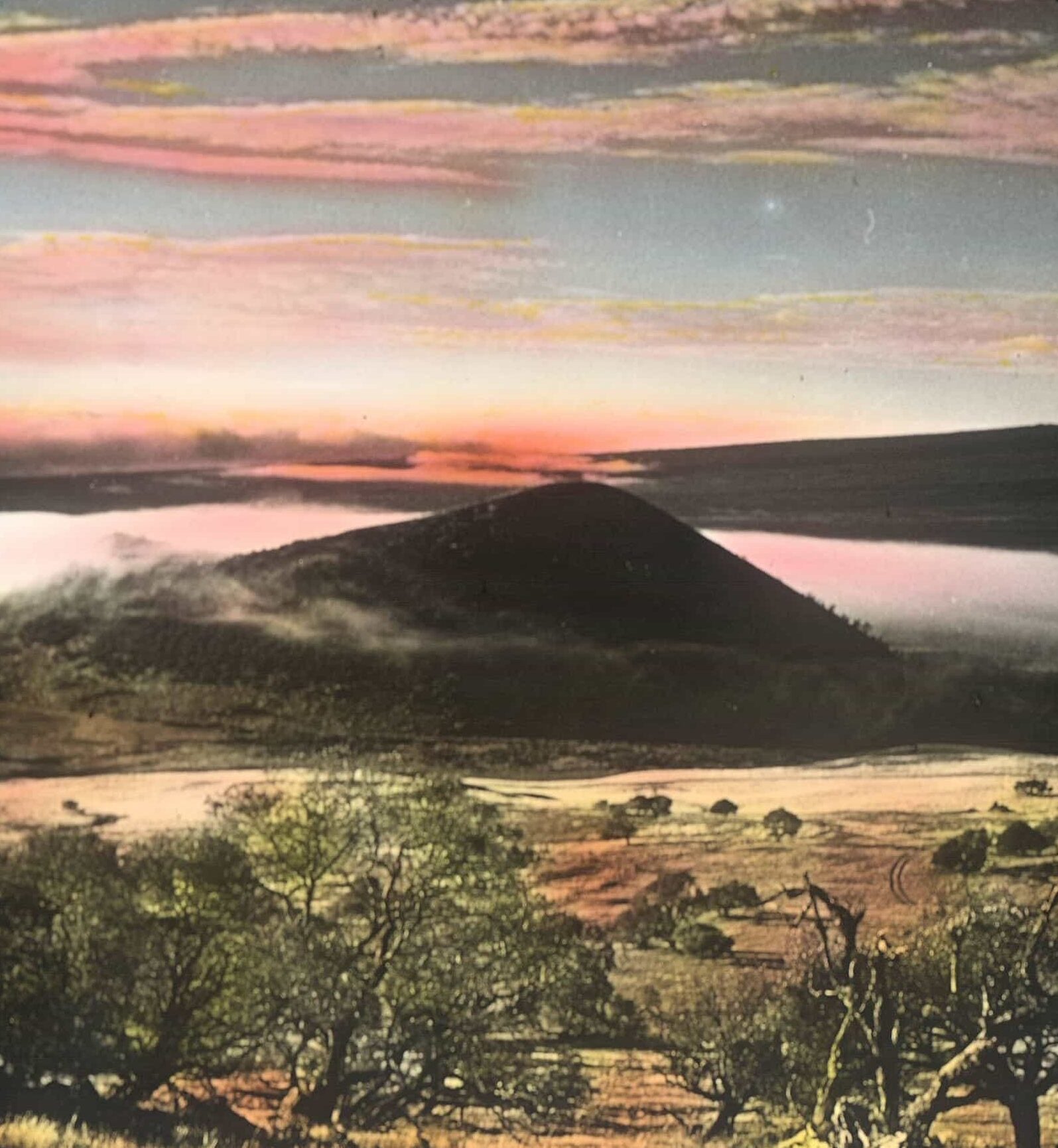 Lantern slide of Mauna Kea during sunrise surrounded by a dense fog. Sparse foliage covers the slopes and clouds are scattered in the sky.