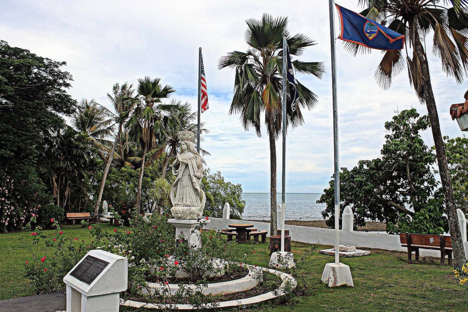 A marble statue of Santa Marian Kamalen, who stands with crown atop her head, in park near ocean. Beside the statue are flags raised on flagpoles.