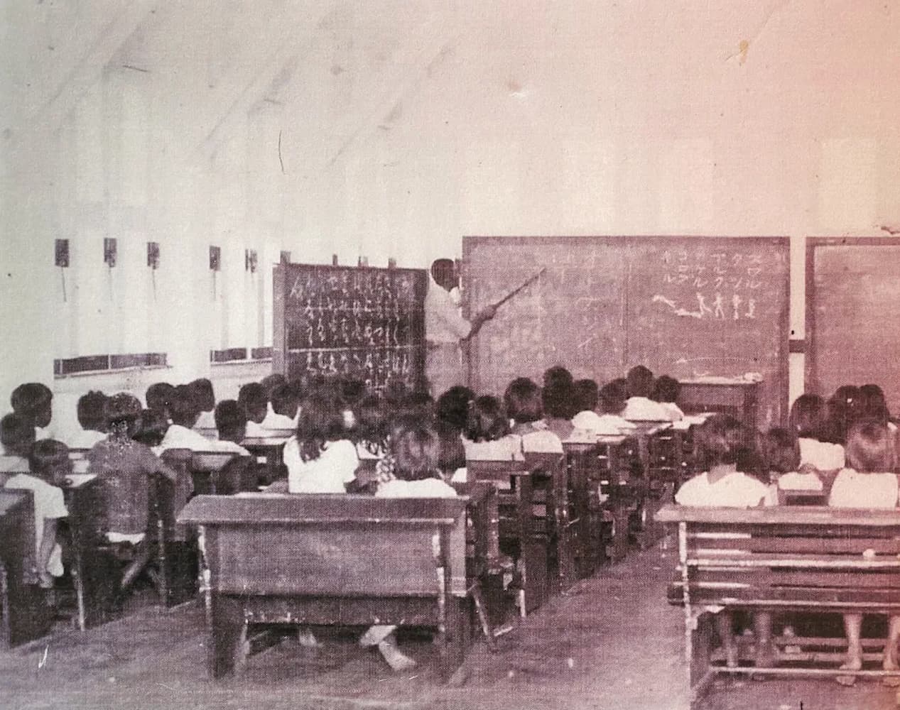 CHamoru students sit in one room schoolhouse. Their teacher, standing near three chalk boards, points to text written in Japanese.