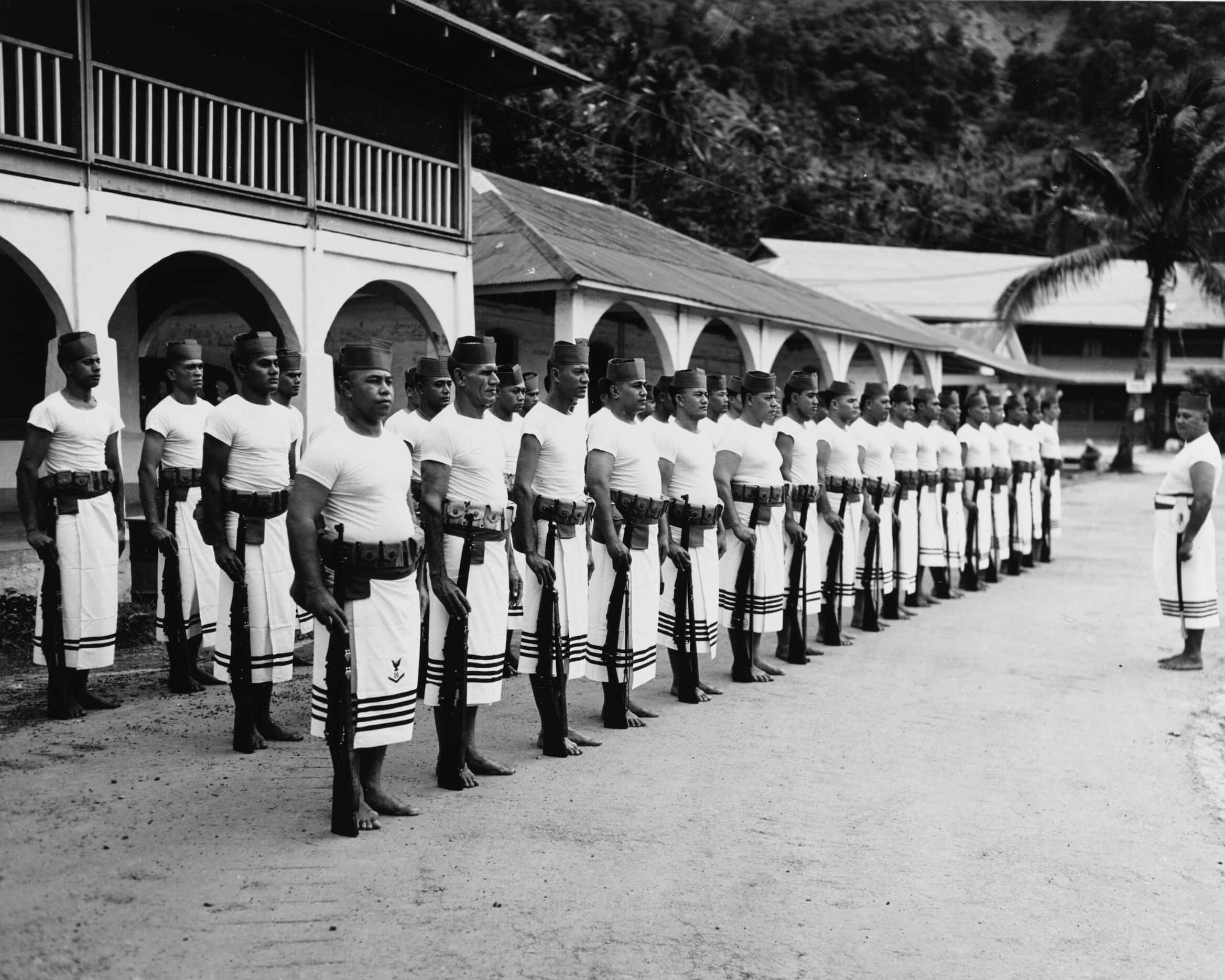 Three rows of Fita Fita guards stand at attention. They hold rifles on a dirt path facing one guard. Buildings and a steep mountain stand behind them.