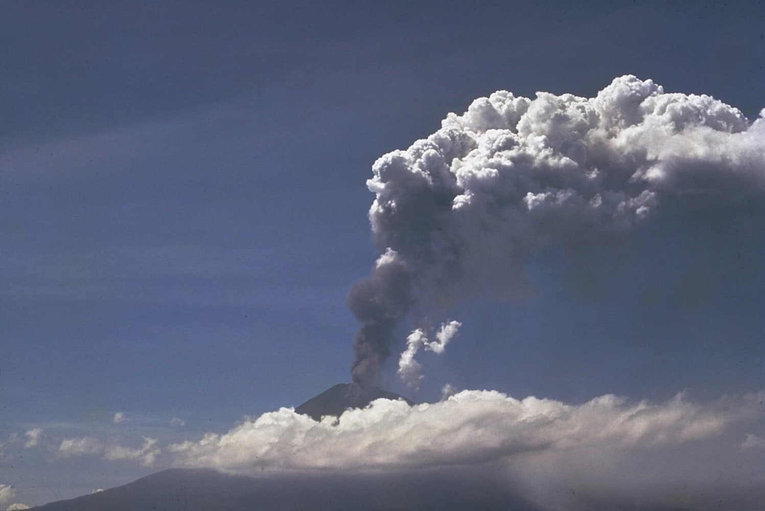 Daytime look at the volcano Ulawun. It hides behind a cloud as the vent at its summit spews out an ash plume into the sky.