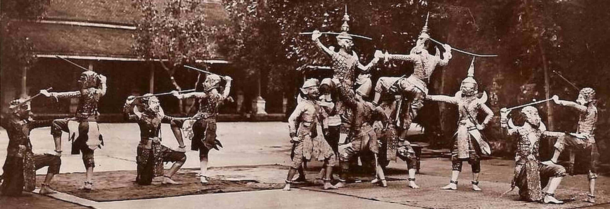 Reamker performance in a concrete courtyard lined with trees. Dancers wear brocaded fabrics and masks and carry sticks in their raised hands.