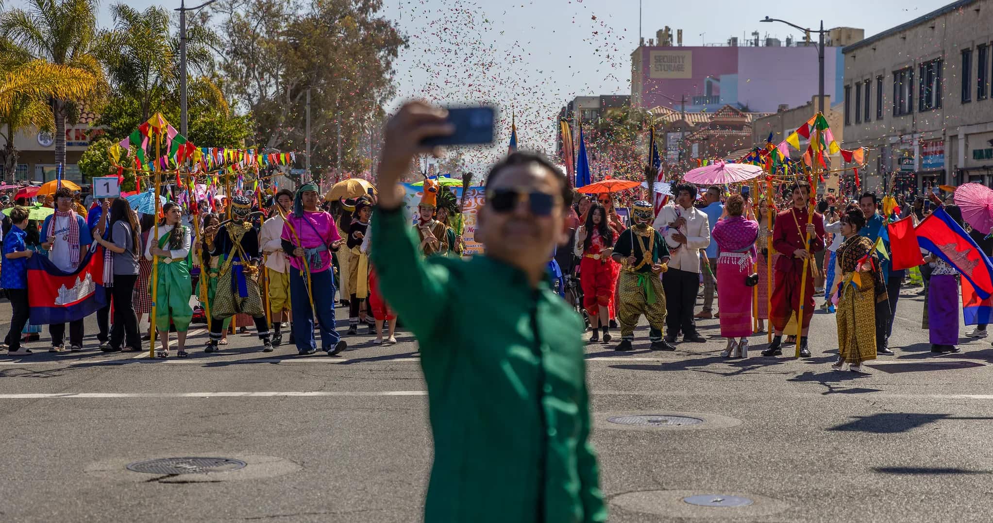 A man takes a selfie in front of the parade in Cambodia Town. Confetti fills the air above the paraders dressed in brightly colored attire.