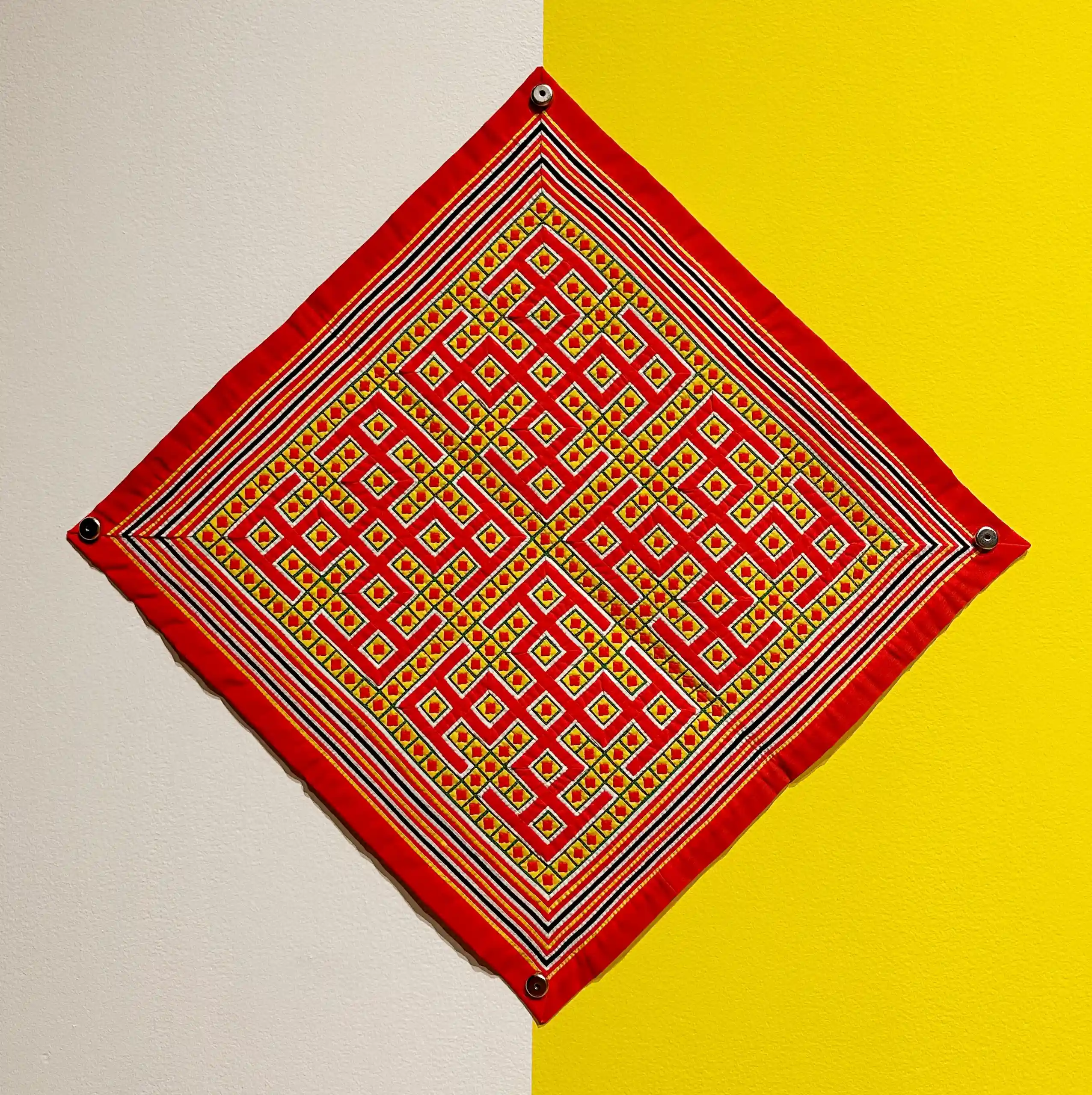 A Noob Ncoos, a diamond shaped piece of red fabric with gold pattern, is photographed against white and yellow backdrop.