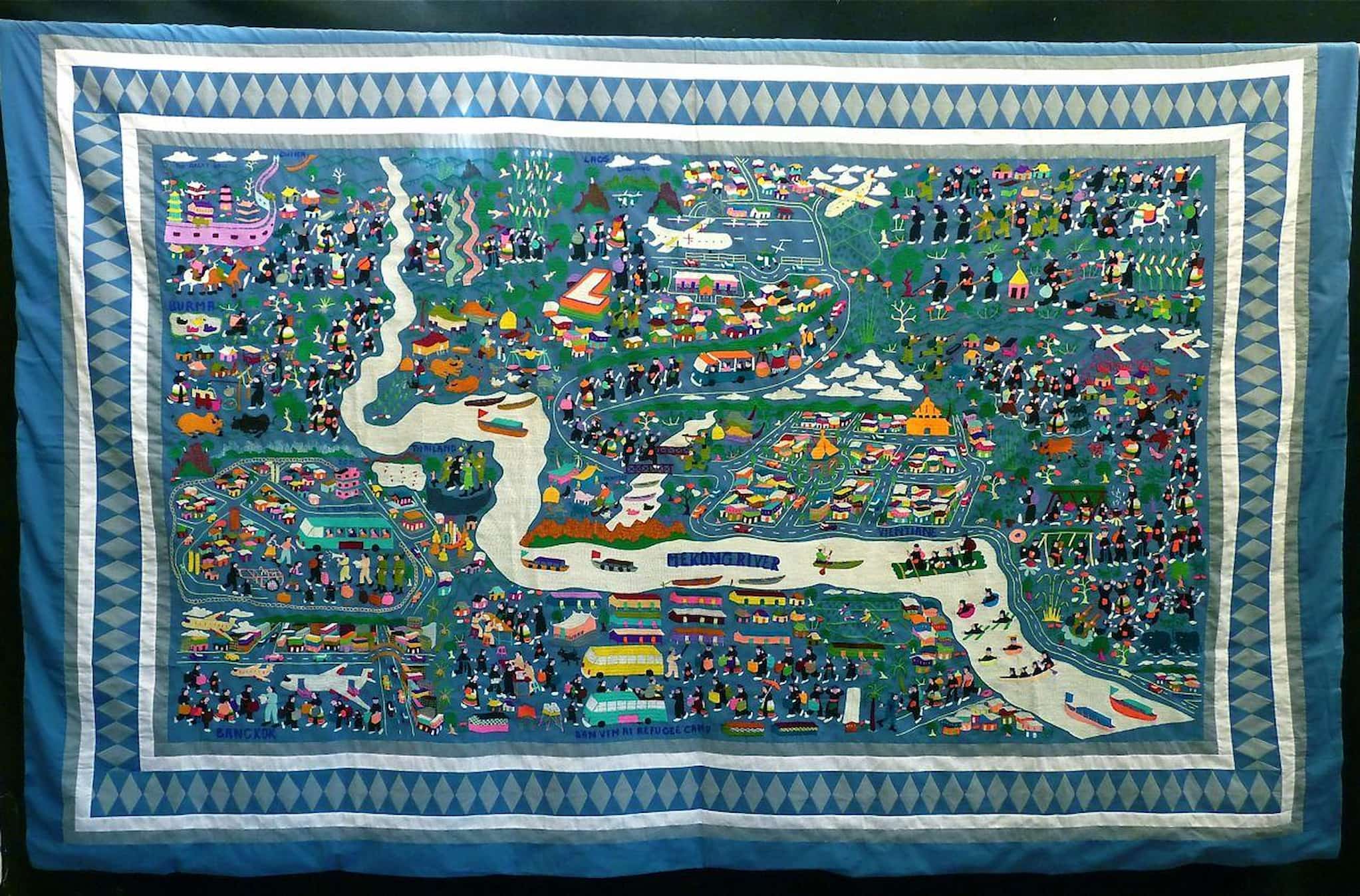 Set against gray fabric, this detailed story cloth shows Hmong people as they walk through lands with colorful buildings bisected by long river.