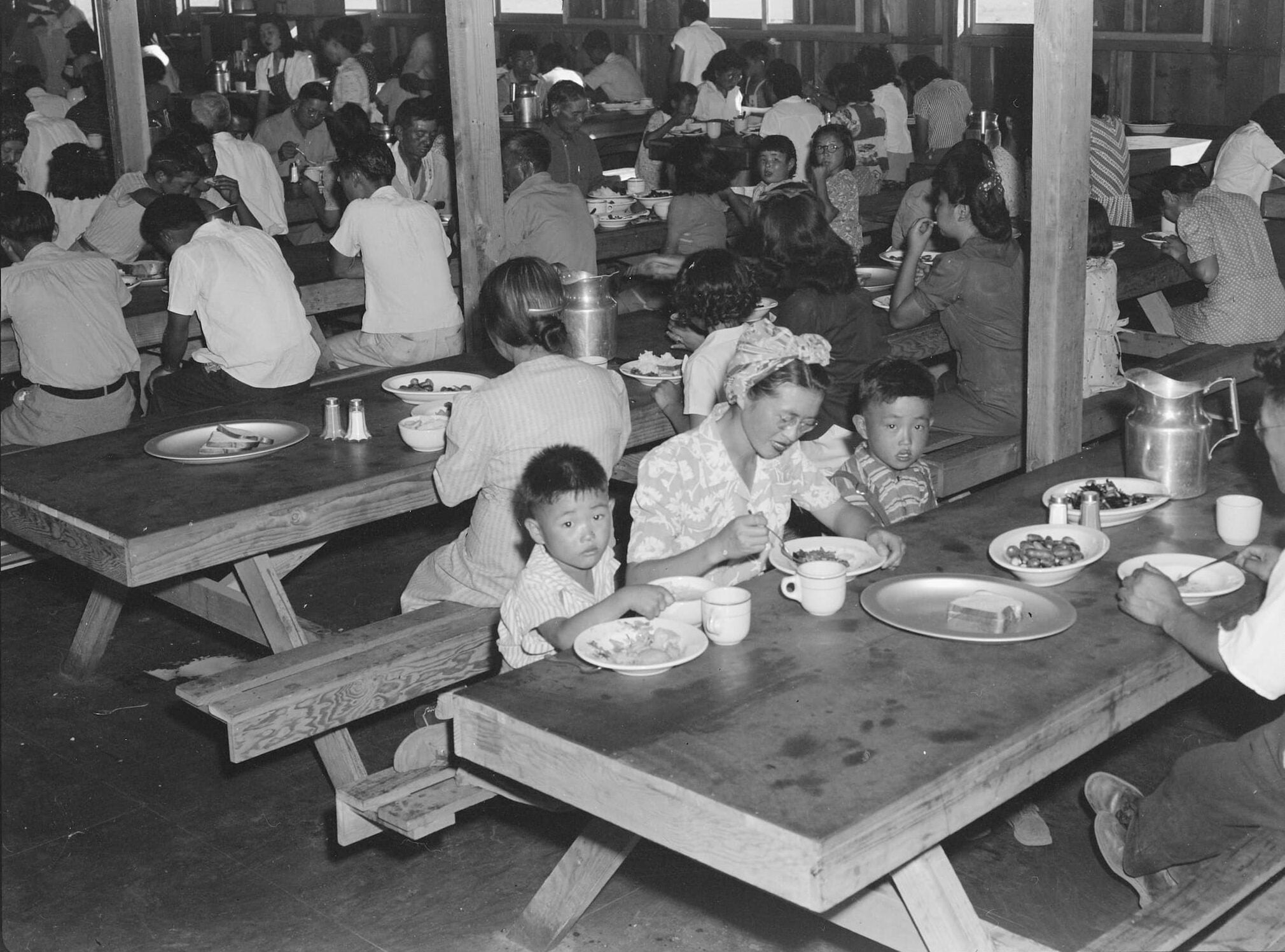 Japanese Americans are seated at picnic tables inside crowded mess hall for meal time.