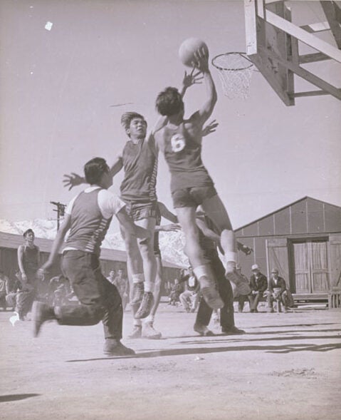 Japanese American men play basketball on outdoor court. Behind them, onlookers sit on benches outside camp buildings.