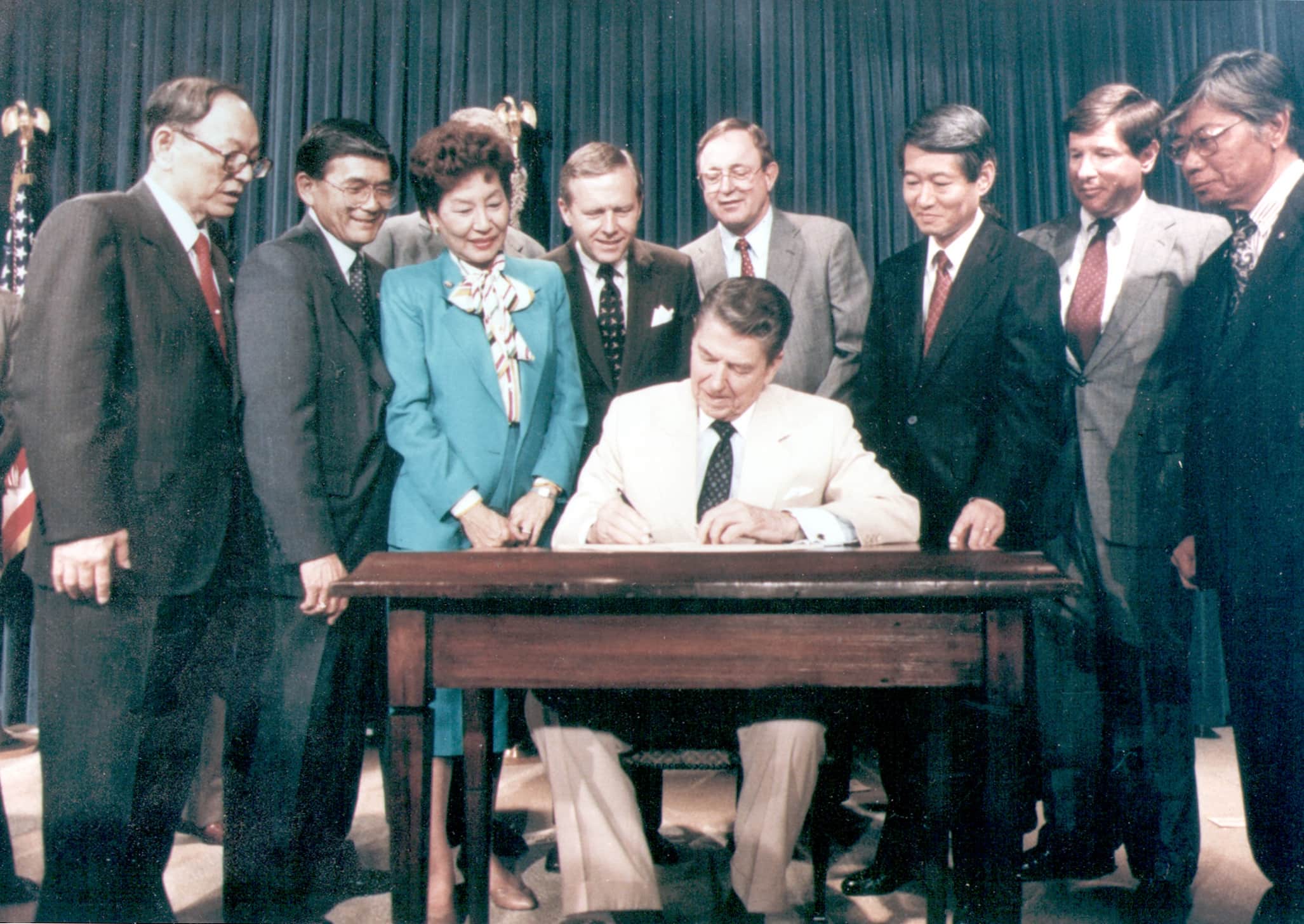 Ronald Reagan, in a white suit, signs paper while seated at desk. Surrounding him are onlookers who stand in half-circle.