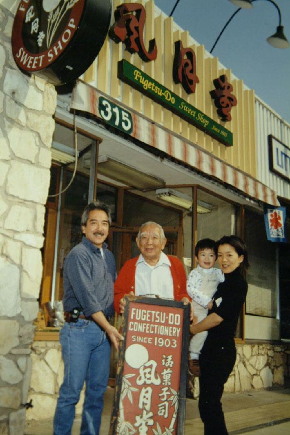 Members of the Kito family, two Japanese American men and a woman carrying a young child, stands on the sidewalk in front of Fugetsu-Do.