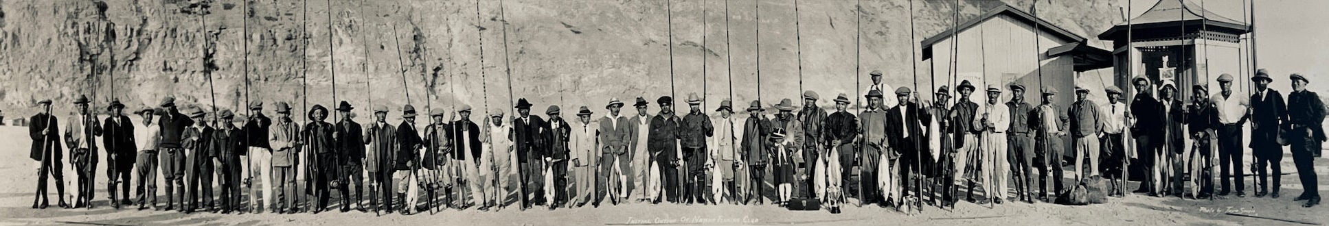 Forty-four Japanese immigrants stand side-by-side, holding fishing poles, as they pose for group photo.