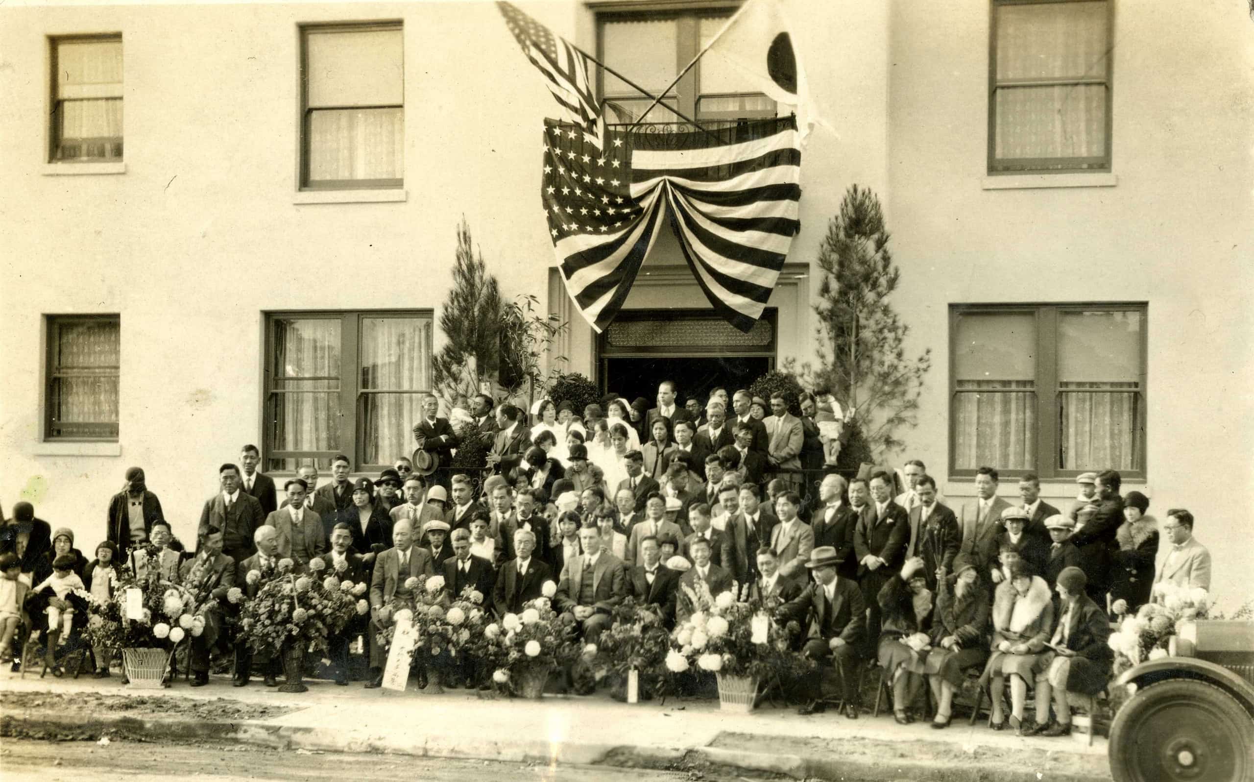 Japanese immigrants gather for group photo in front of the Japanese Hospital of Los Angeles. Above the entryway, the flags of the U.S. and Japan fly.