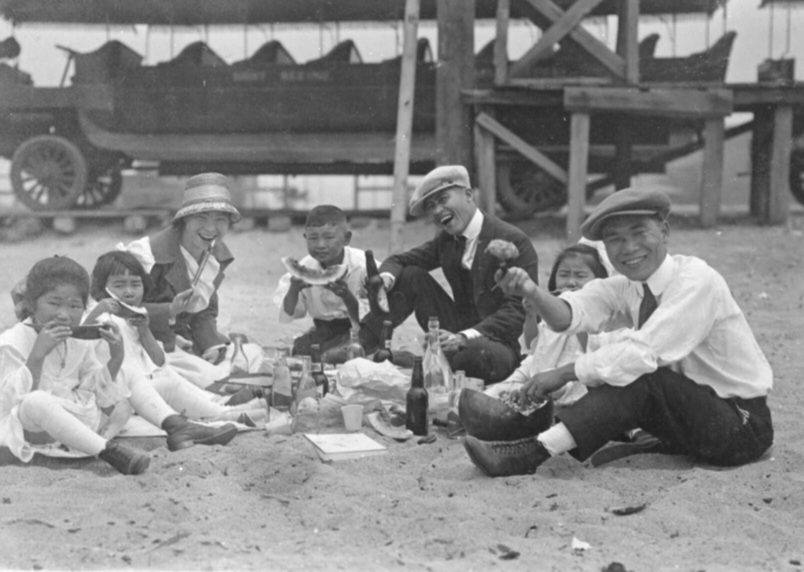 A Japanese American family sits on the sand while eating watermelon. An empty carriage with nine rows is standing in the background.