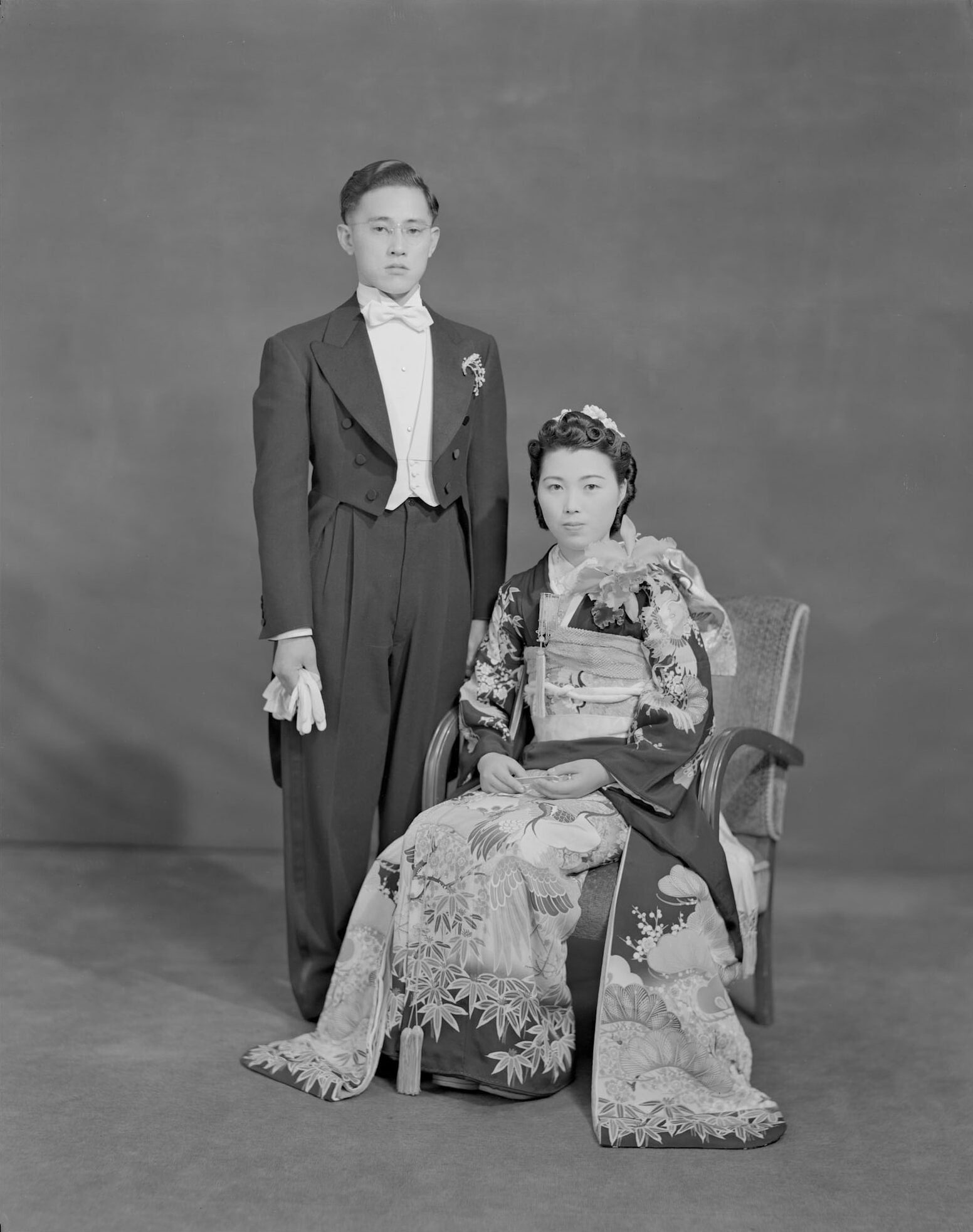 A Japanese American wedding photo with a dark background. The husband stands on the left in a tuxedo and the wife sits on the right in a kimono.