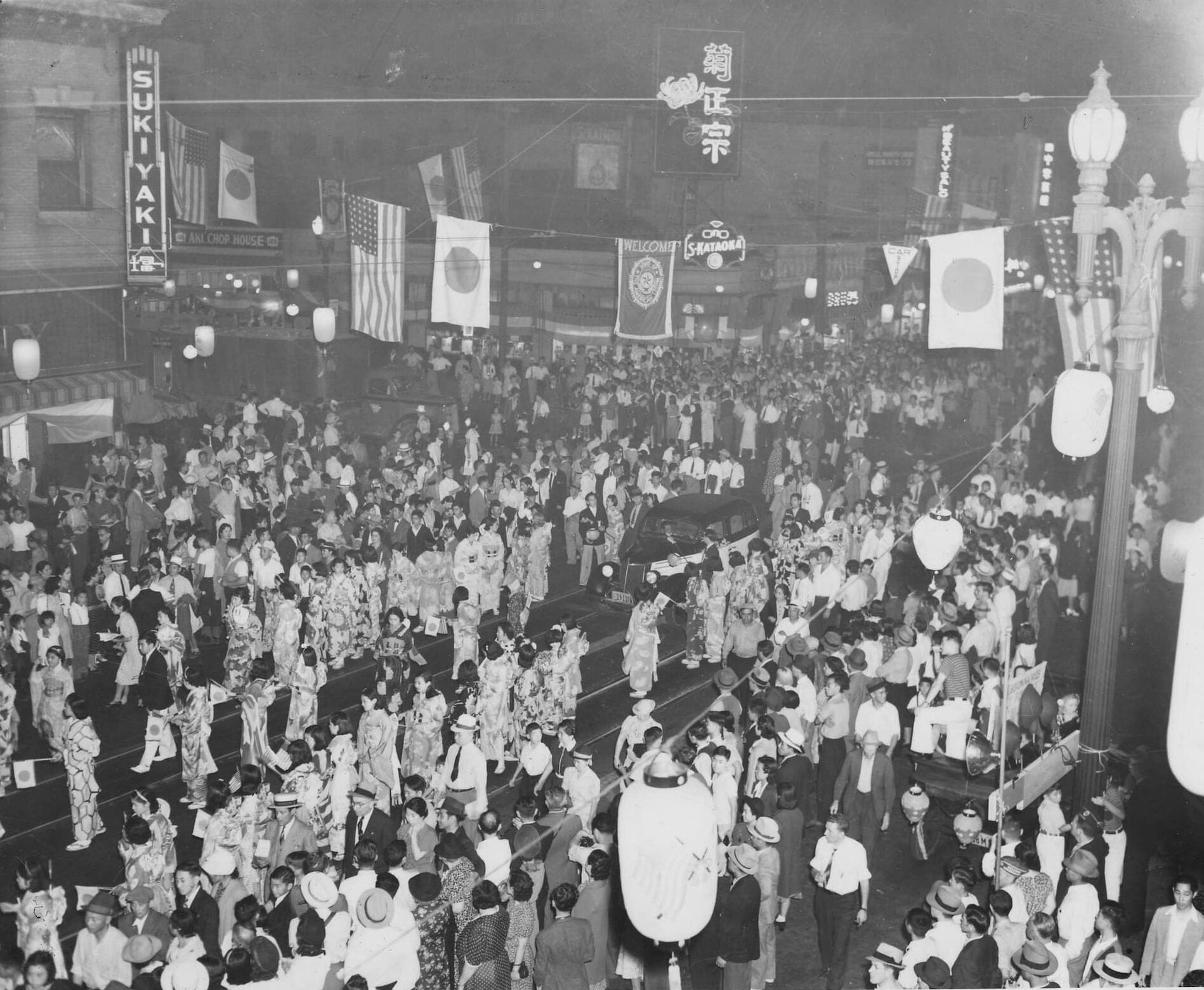 Large crowds of White and Japanese Americans line the streets. The flags of U.S. and Japan hang overhead along lamp posts.