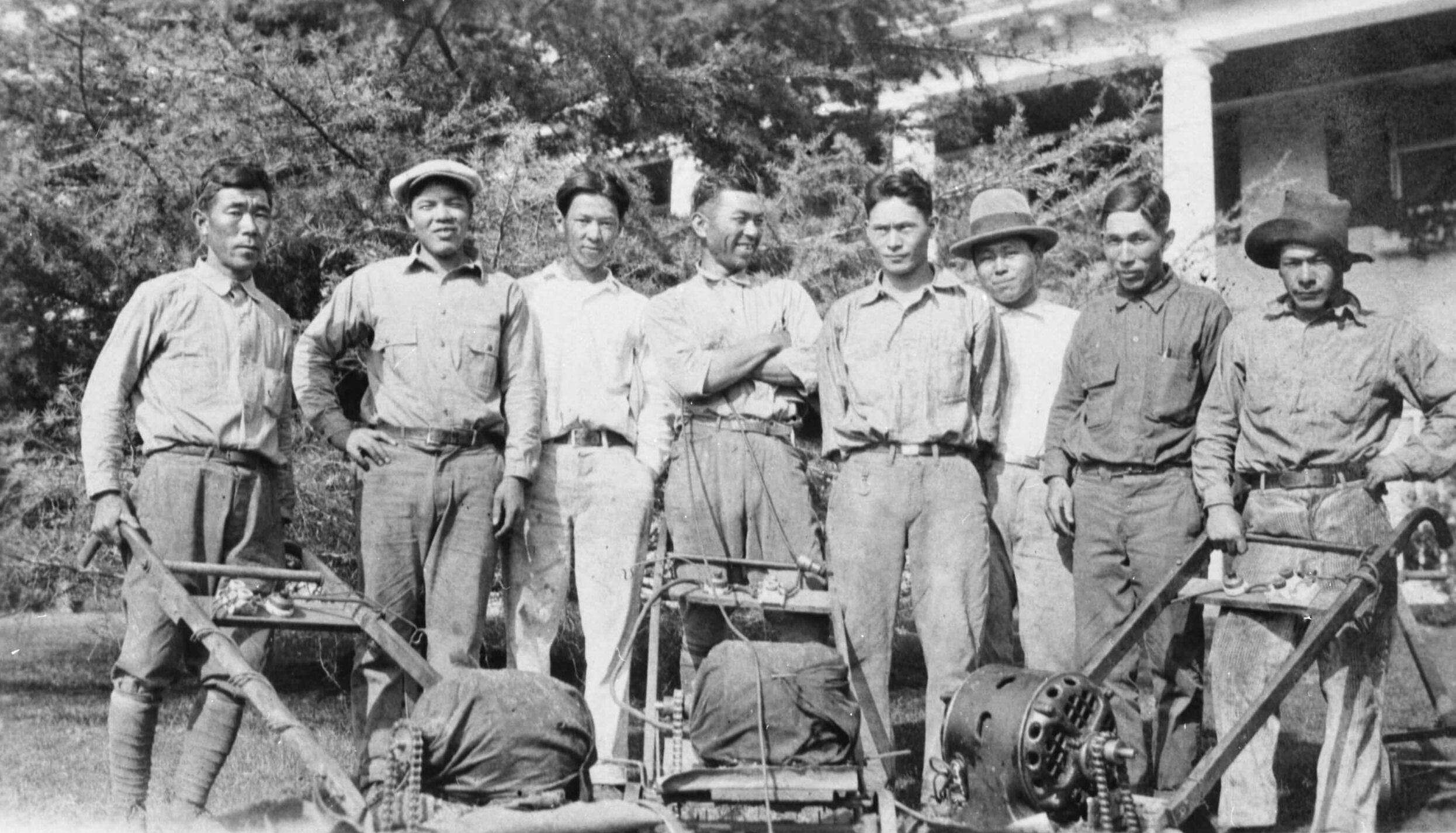 Eight Japanese Americans stand together in front of a house. Three lawn mowers sit in front of them.