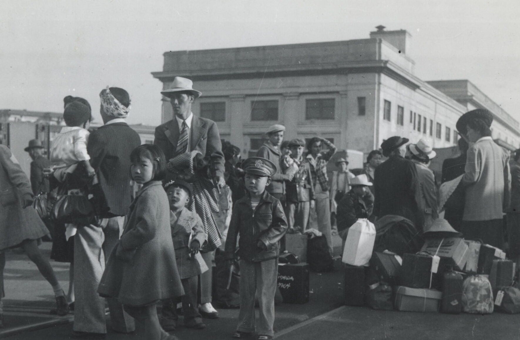 Groups of Japanese American families dressed in Sunday suits and coats wait outside to board trains with their luggage piled beside them.
