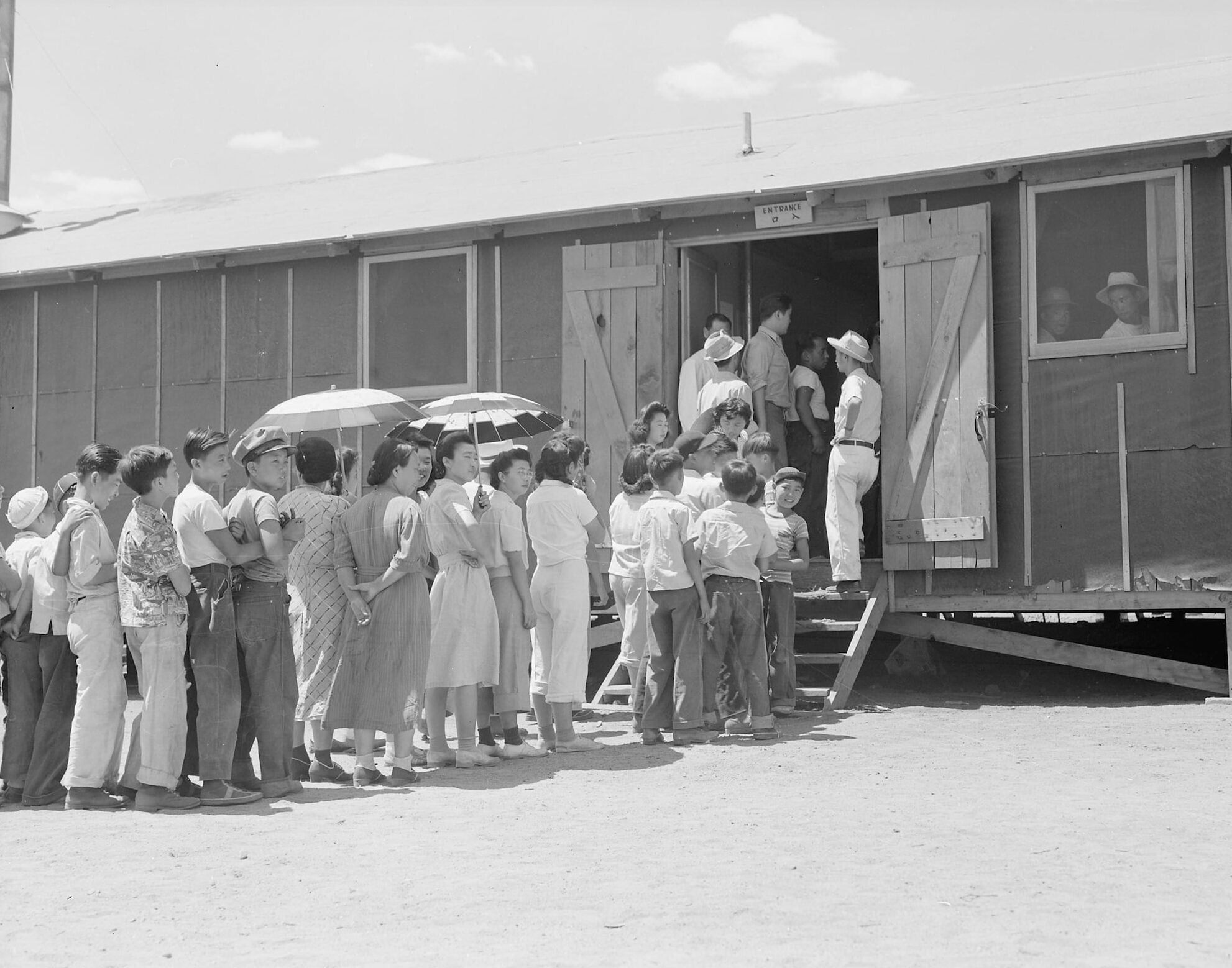 A line of Japanese American adults and children form line as they wait outside the mess hall building.