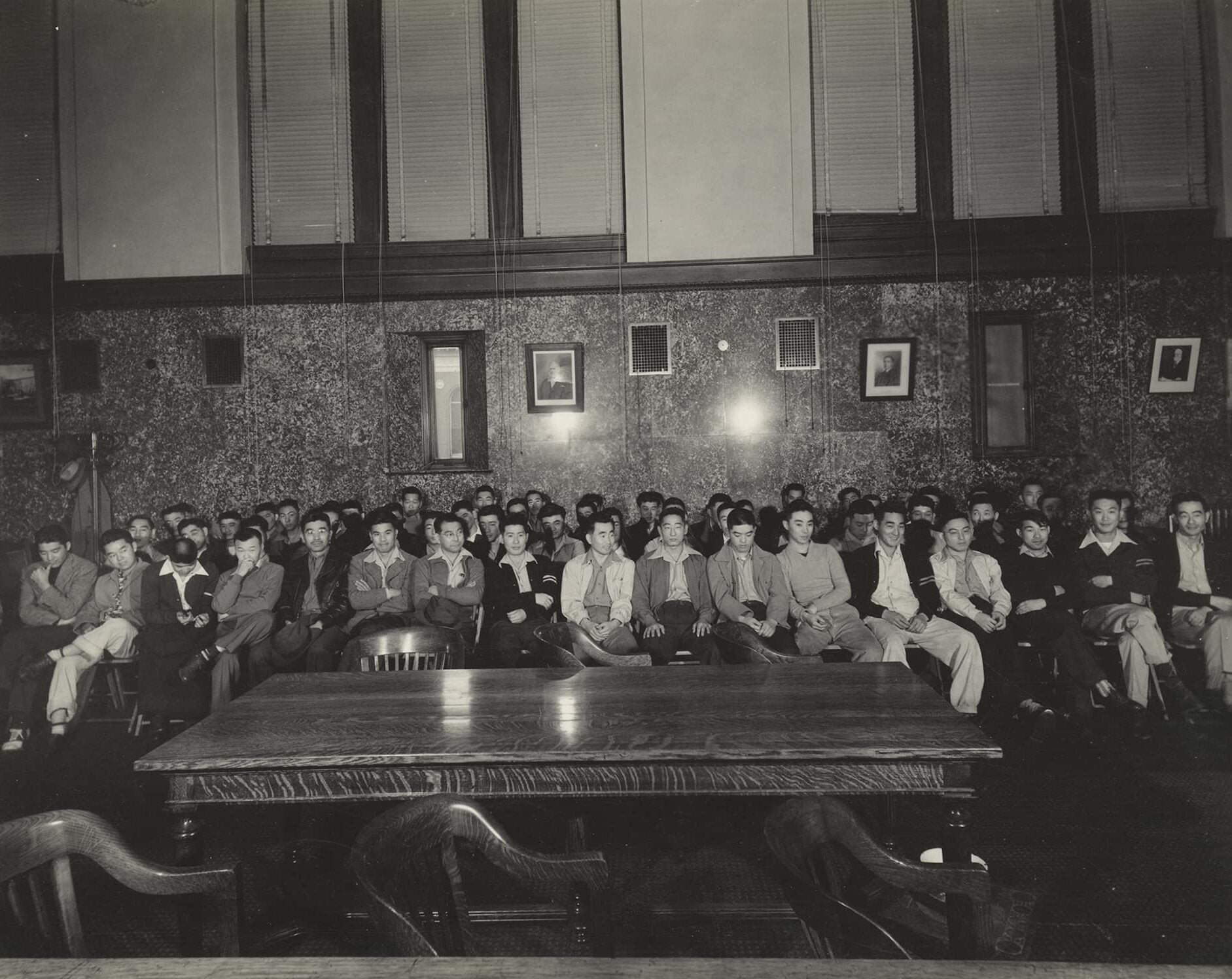 Sixty-three Japanese American men are seated in rows within a large courtroom. Along back wall are framed portraits and windows with shades drawn.
