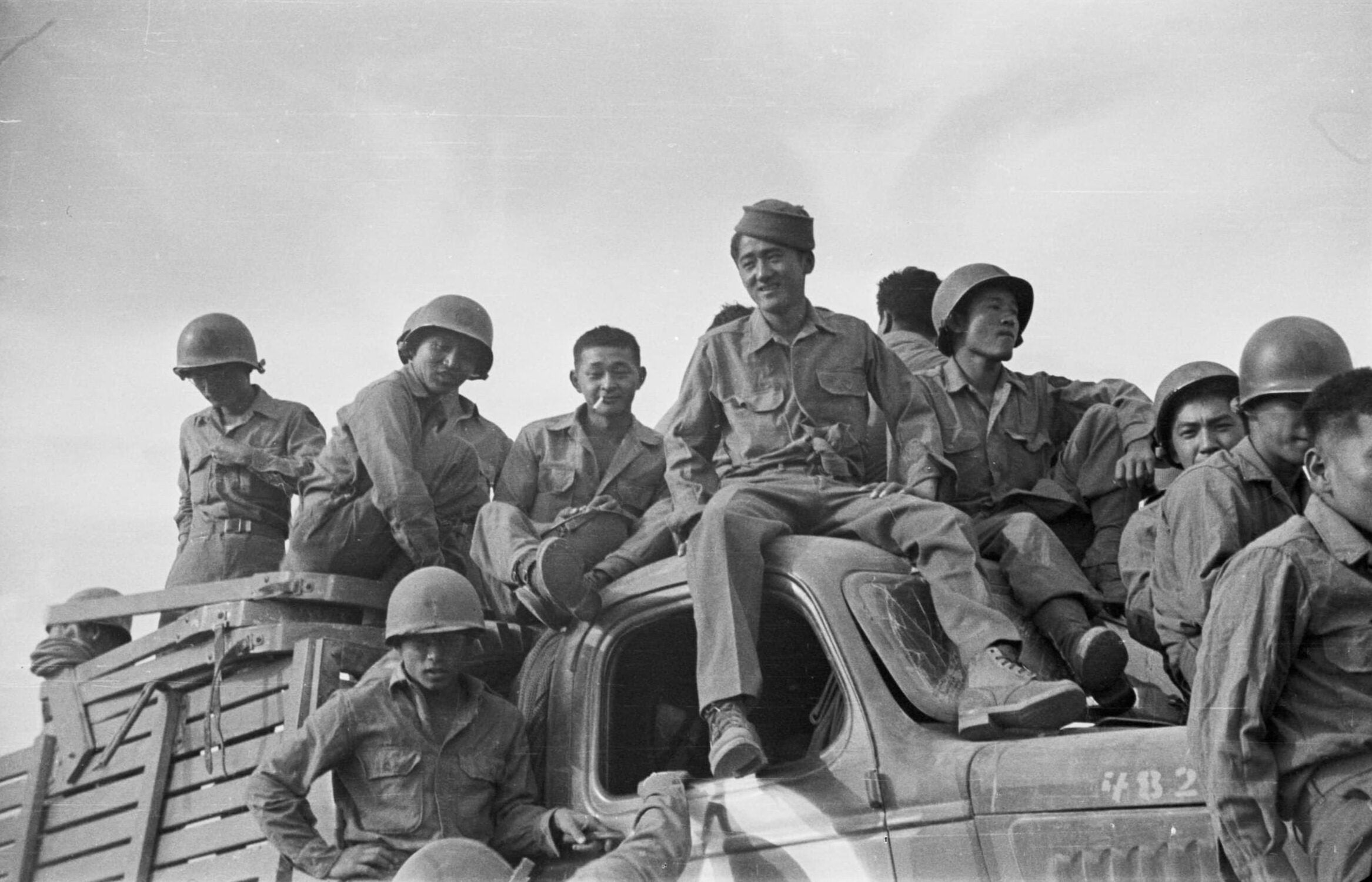 Twelve Japanese American soldiers, dressed in uniform, sit along roof and on hood of a truck.