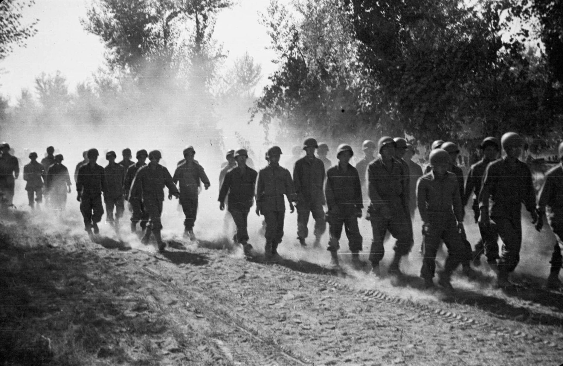 Uniformed Japanese American soldiers march through outdoor clearing, with dust and dirt being kicked up in the air.