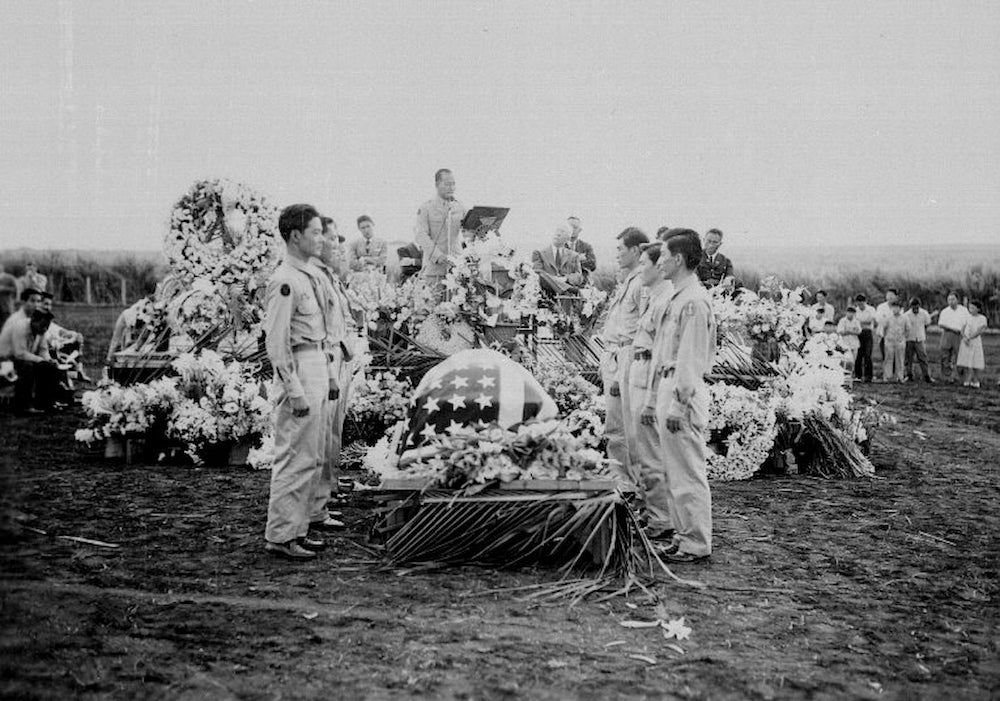 Soldiers stand around a coffin, draped in U.S. flag, during outdoor funeral. Men in suits sit on platform, while man stands at center podium.