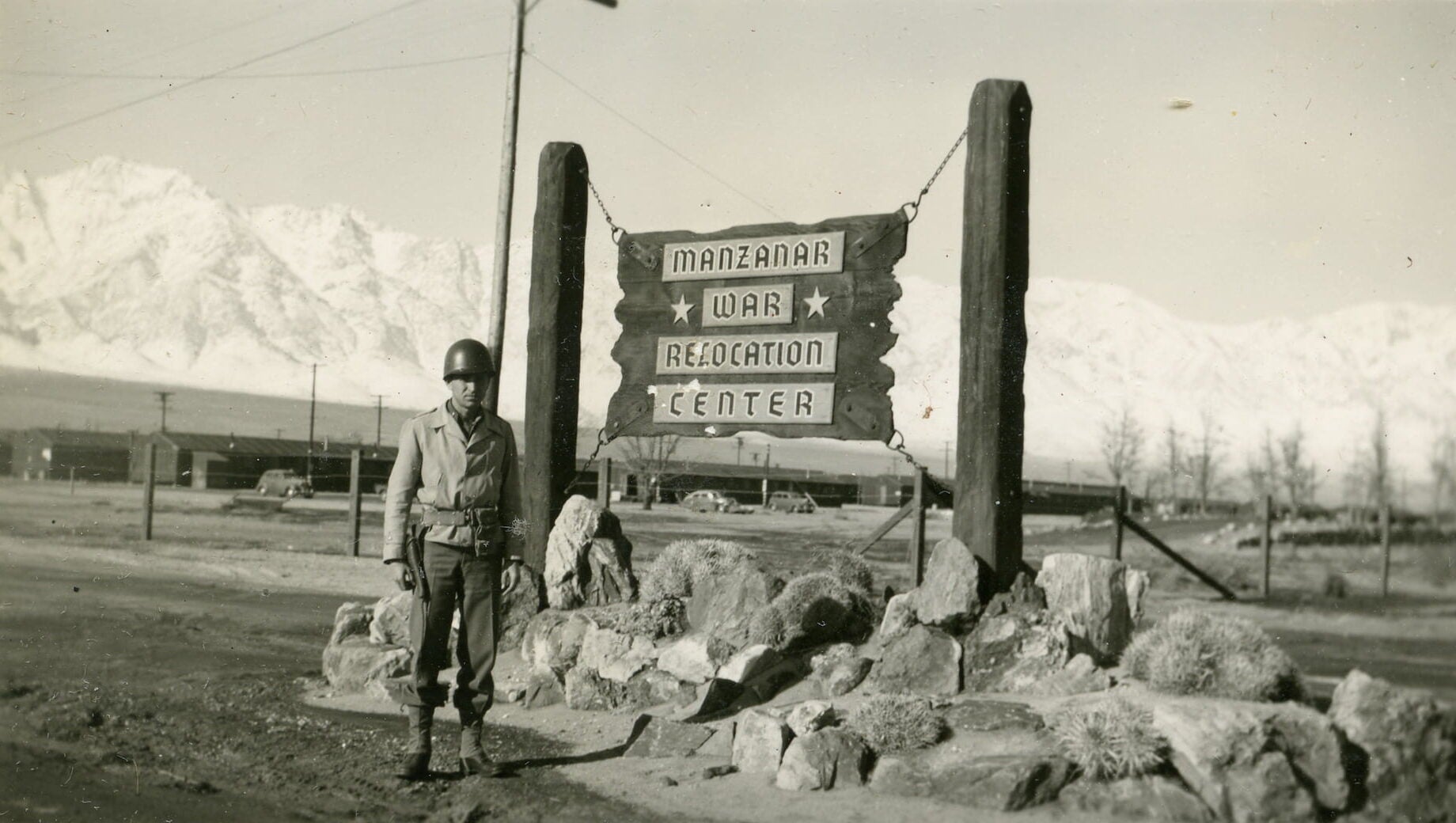 A soldier stands in front of a sign reading, "Manzanar War Relocation Center." The sign sits atop rocks and mountains cover the background.