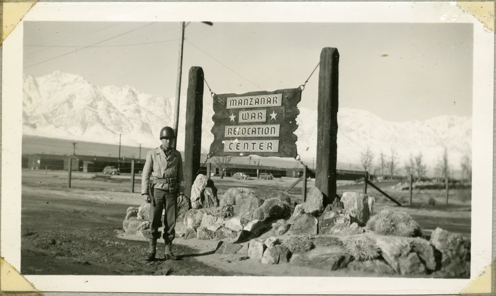 A soldier stands in front of a sign reading, "Manzanar War Relocation Center." The sign sits atop rocks and mountains cover the background.