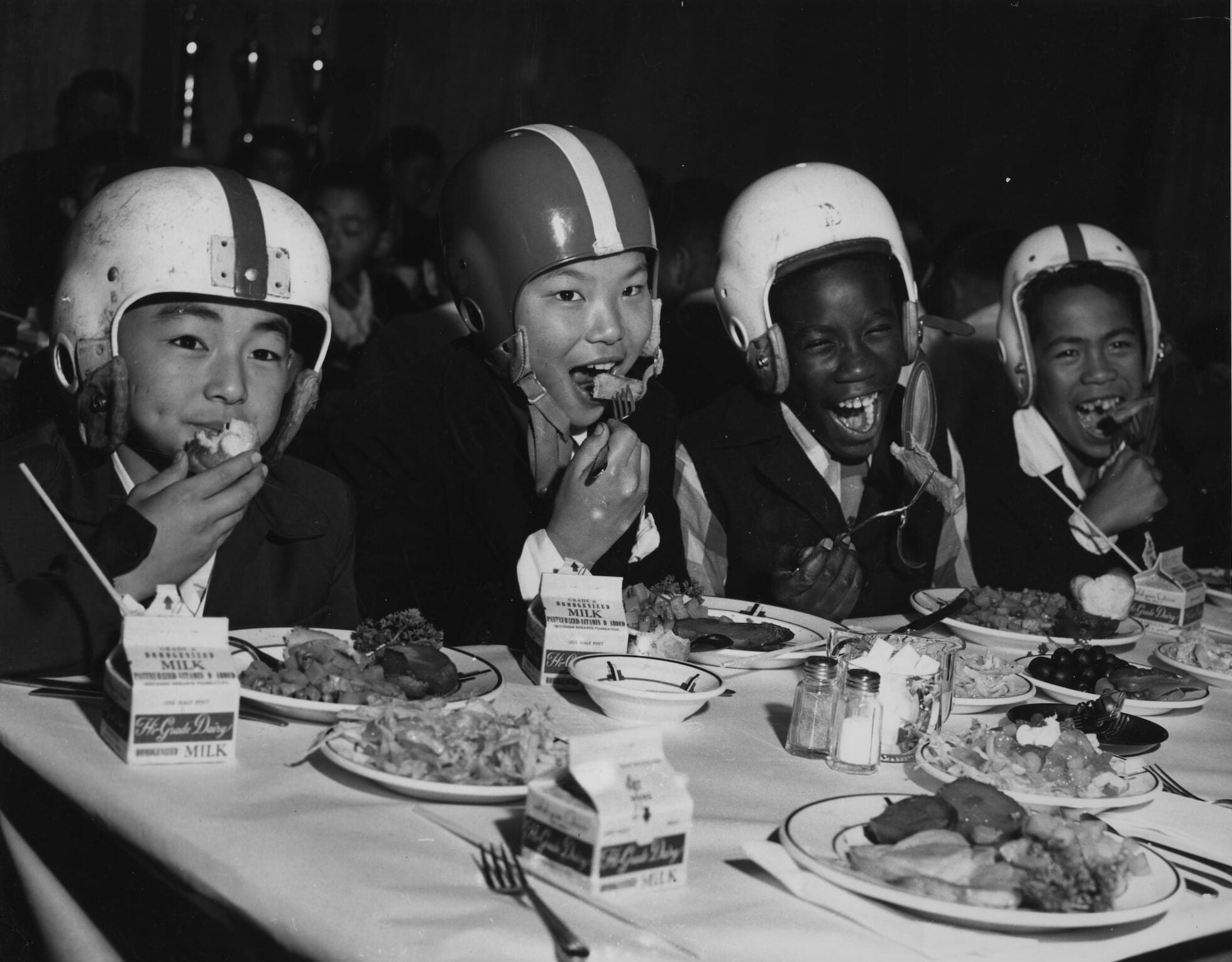 Two Japanese American kids on the left and two African American kids on the right eat together. They are dressed in their football gear.